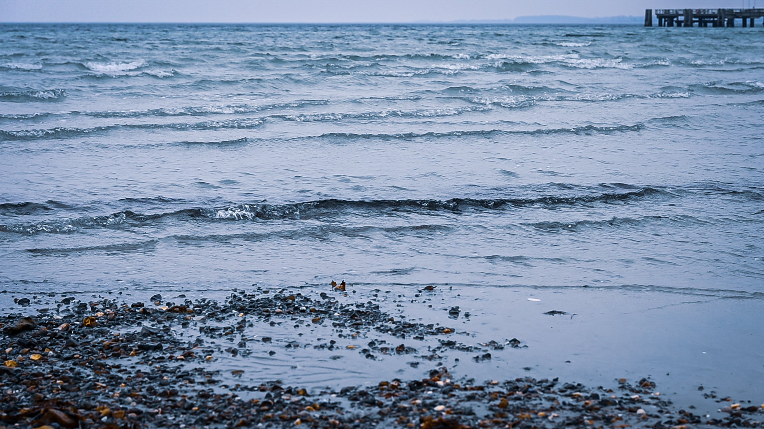 A view of the ocean with small waves and a rocky shoreline, with a pier extending into the water on the right side of the image.