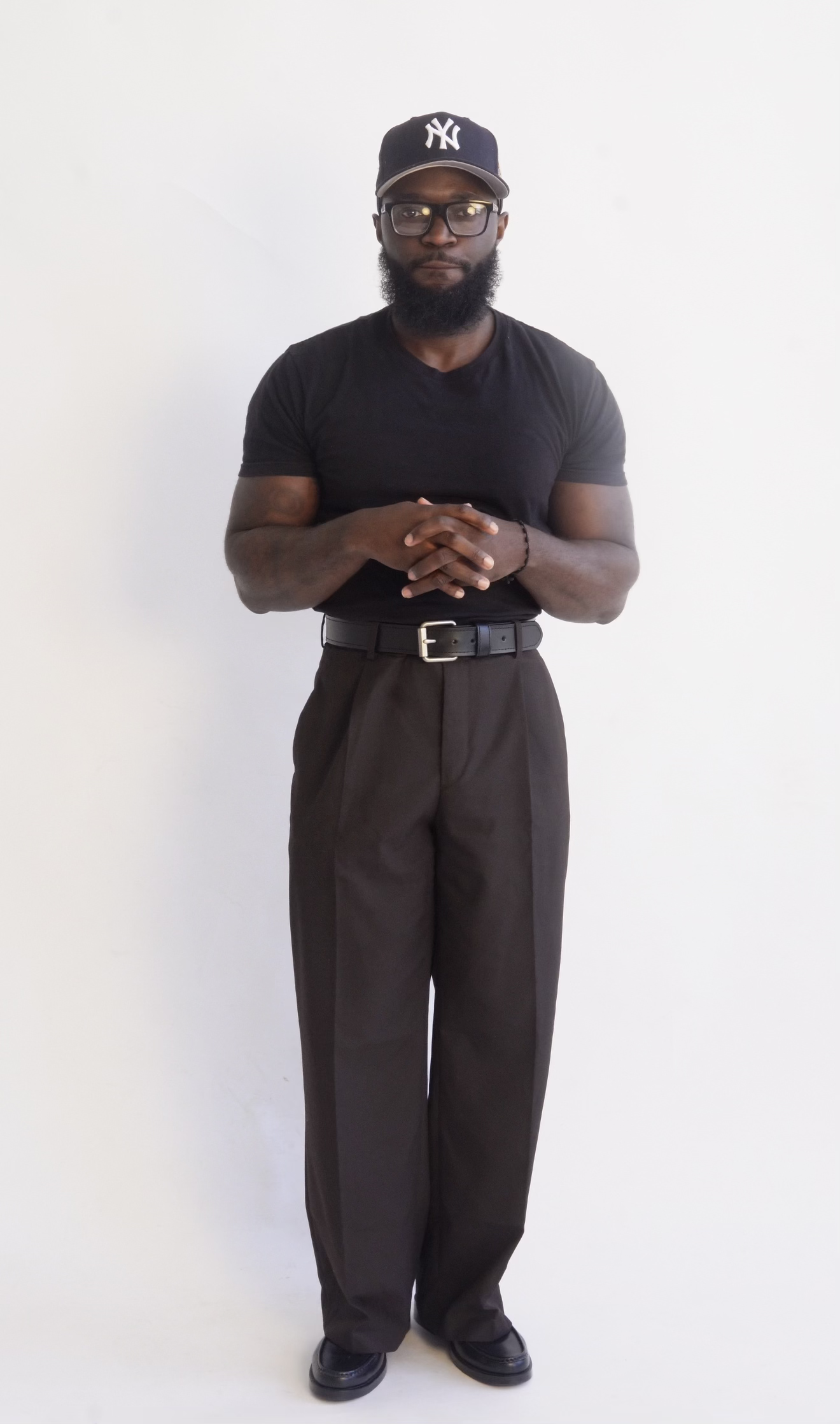 A man with a beard and glasses wearing a black New York Yankees cap, black t-shirt, brown trousers, and black shoes, standing in front of a plain white wall.