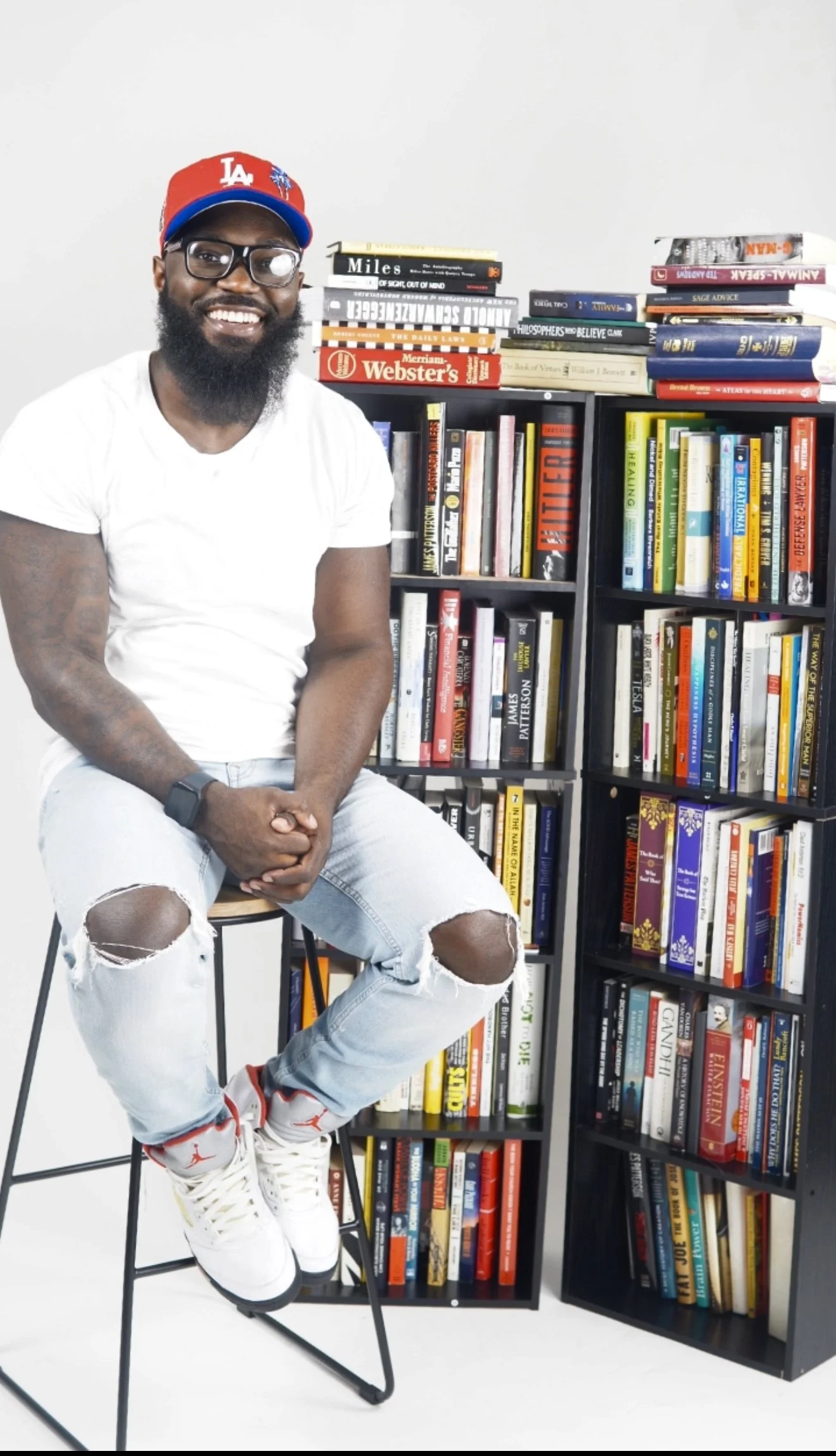 Smiling man sitting on a high stool in front of bookshelves filled with books, wearing glasses, a red baseball cap, a white T-shirt, ripped jeans, and sneakers.