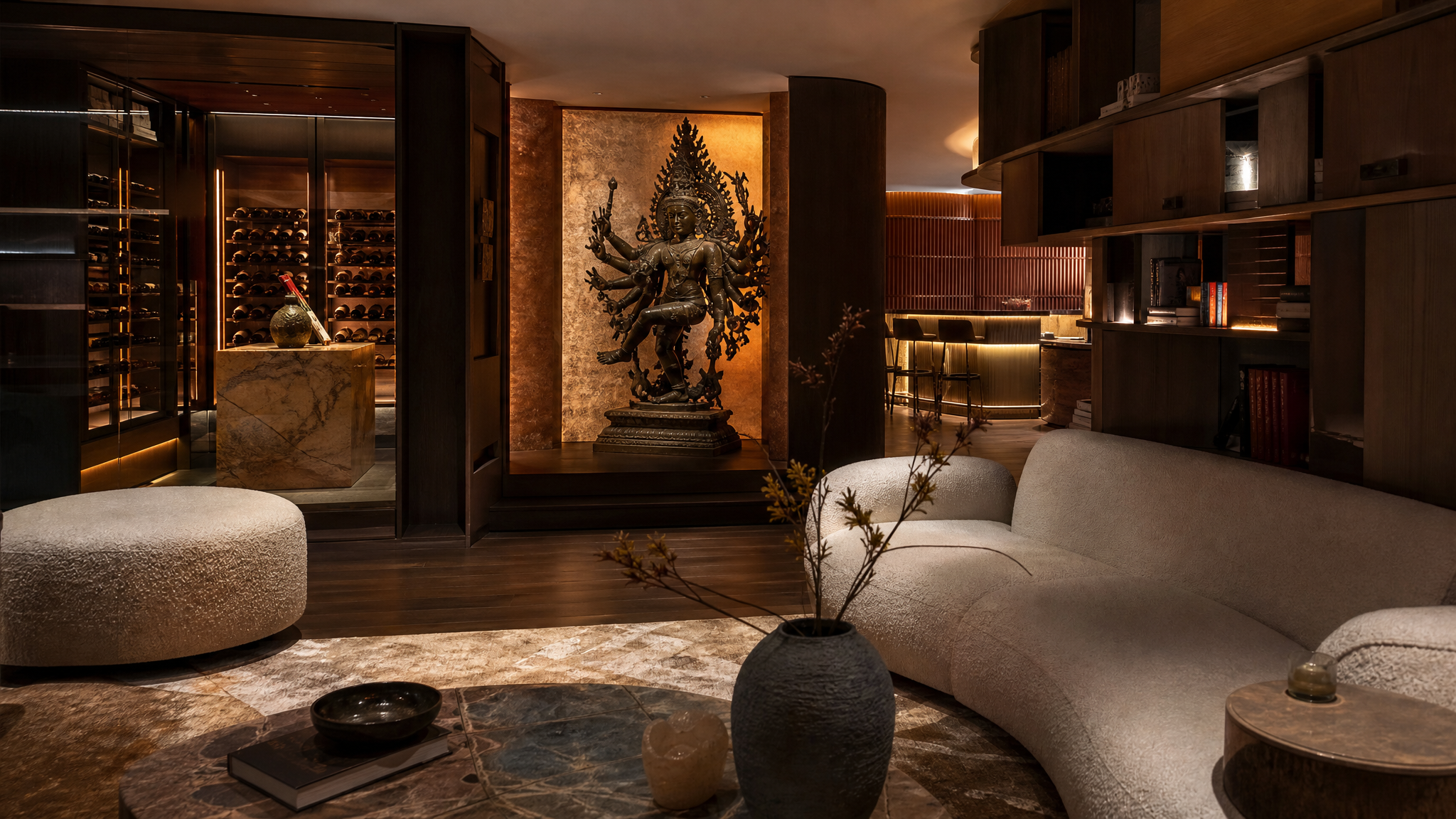 A modern living room with curved beige couches, a marble coffee table, and decorative vases. Behind a wooden partition, a statue of a multi-armed deity is illuminated, with wine storage on the left and a bar area with barstools on the right.