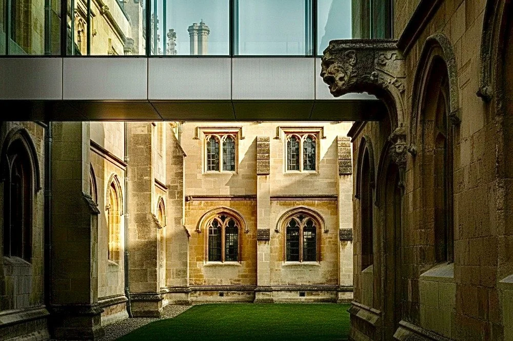 A view of an architectural courtyard between historic stone buildings, with a modern glass bridge connecting them above.