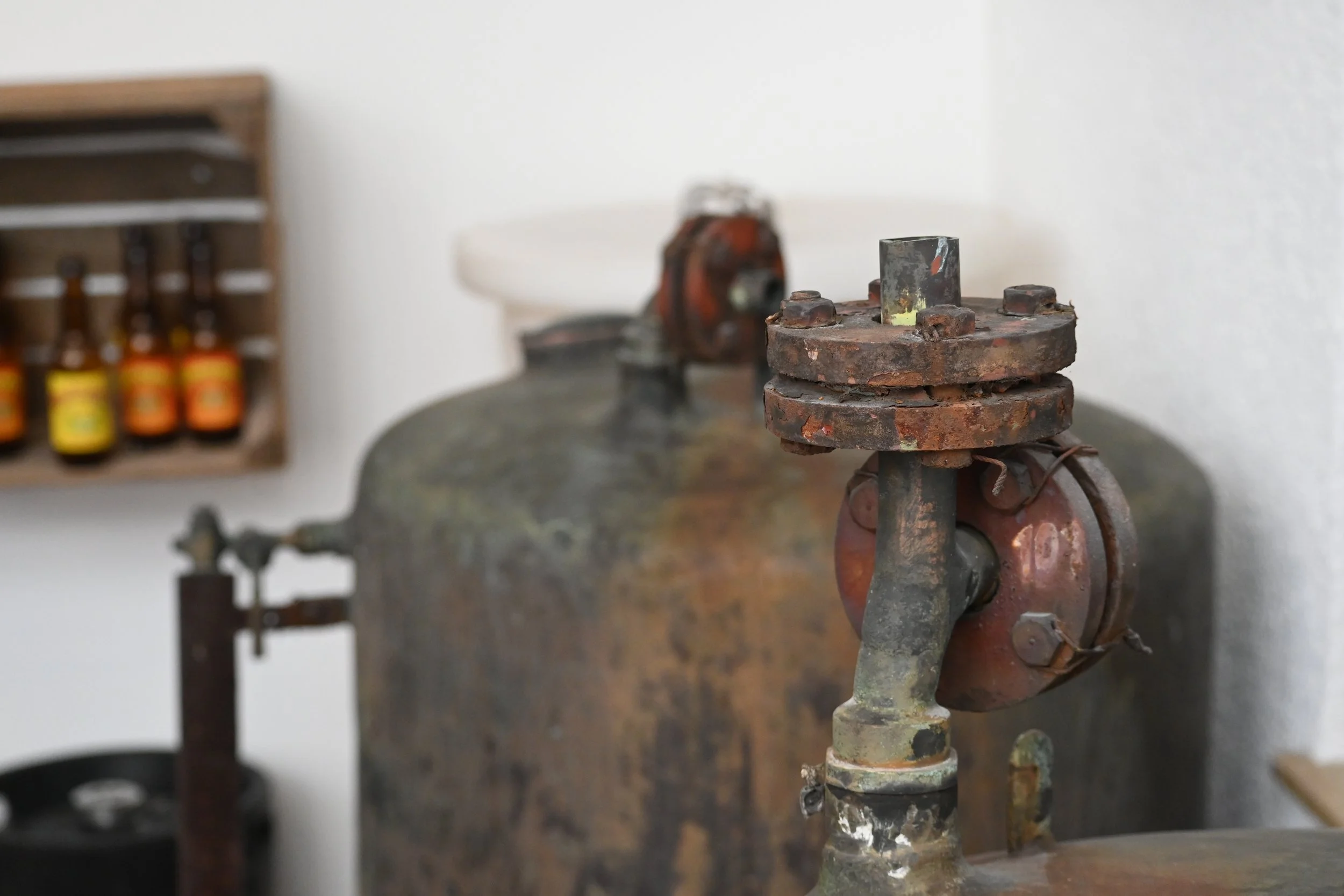 Ein alter, verrosteter metallischer Apparat mit mehreren Teilen und Schrauben, auf einer rustikalen Oberfläche, im Hintergrund eine Wand mit einem Holzregal, das kleine Flaschen mit gelben und orange Etiketten hält.