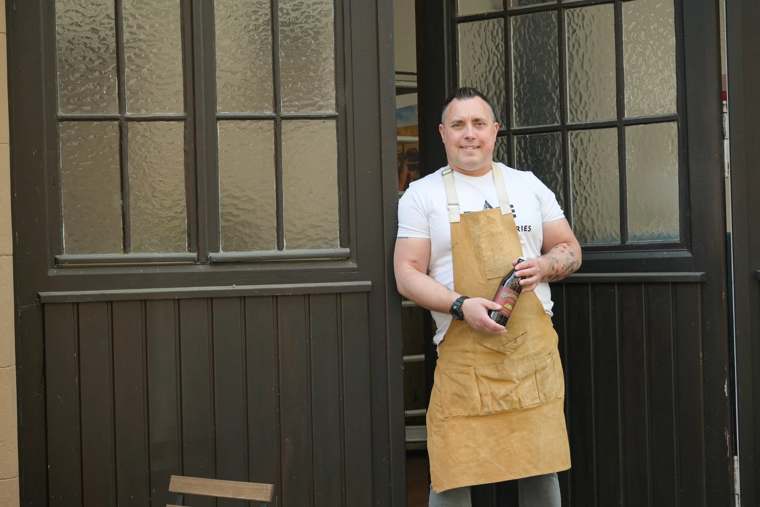 Mann in weißem T-Shirt und brauner Schürze steht vor einer Holztür mit Glasfenstern, hält eine Flasche Bier in der Hand.