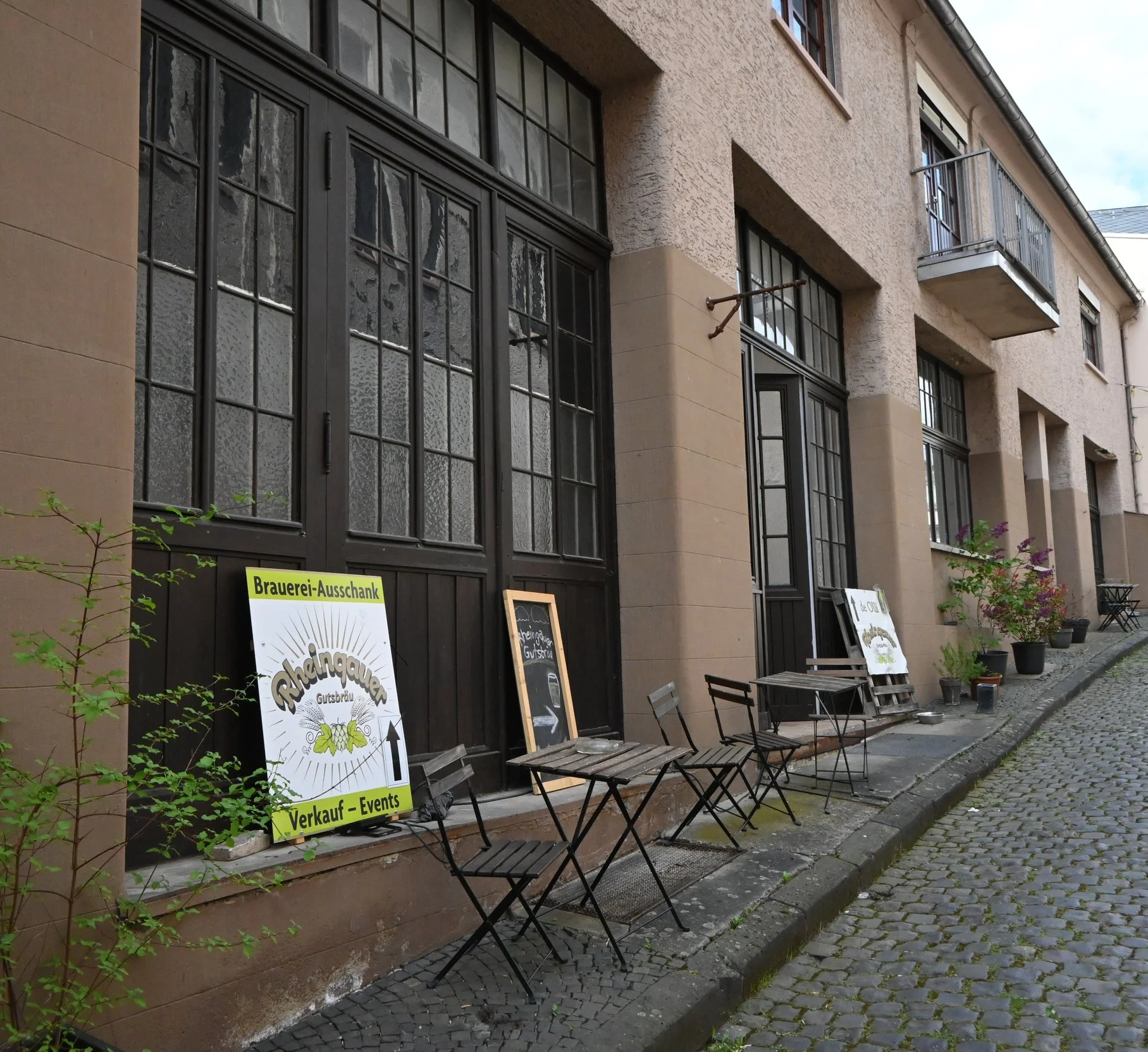Außenansicht eines Gebäudes mit Terrassenmöbeln und Schildern, die Verkostungen und Veranstaltungen ankündigen, sowie Pflanzen in Töpfen.