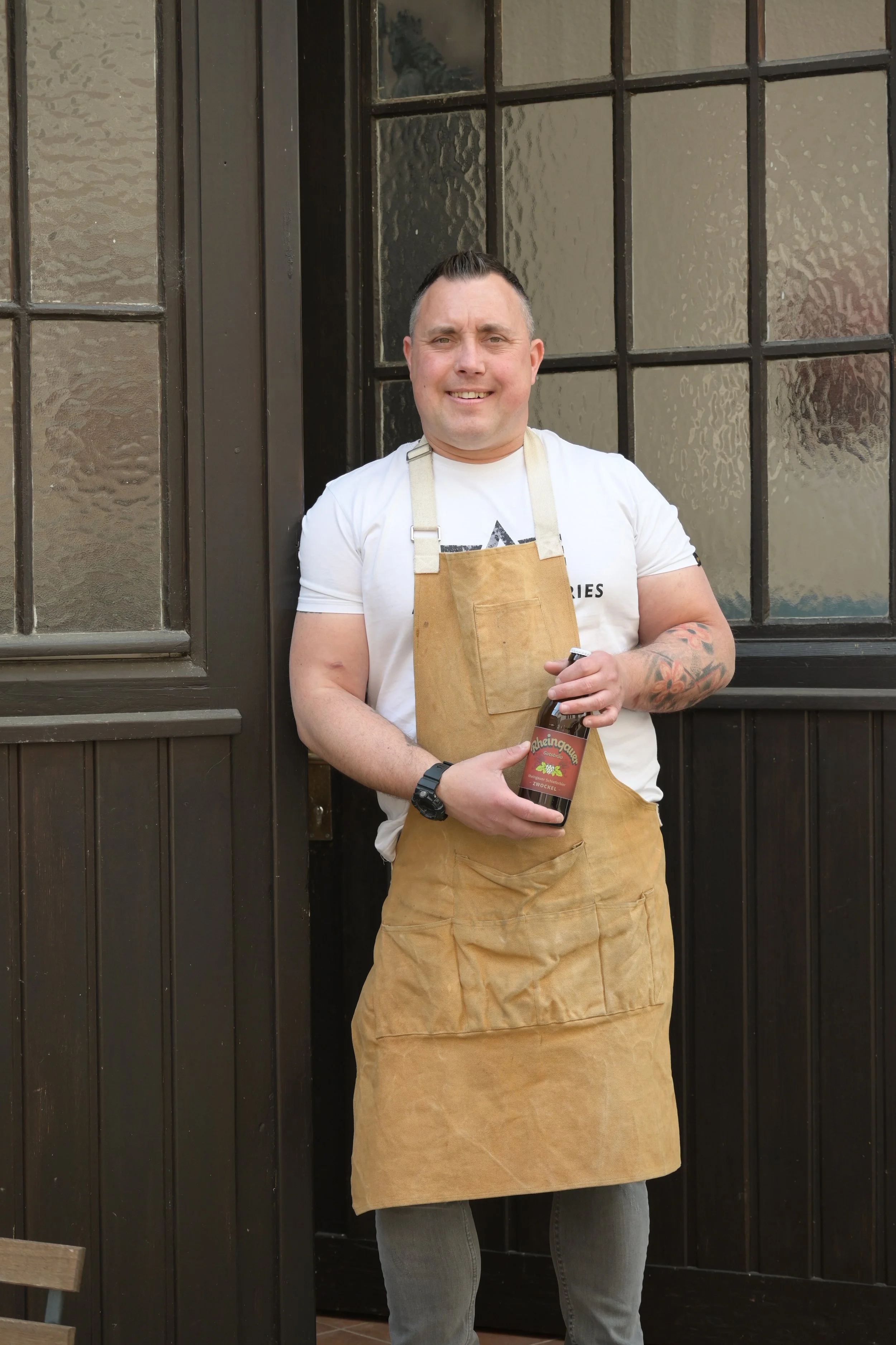 Ein Mann trägt eine braune Schürze, hält ein Flaschen Bier, steht vor einem Fenster mit dunklen Rahmen, trägt ein weißes T-Shirt, hat Tattoos am Arm und lächelt.
