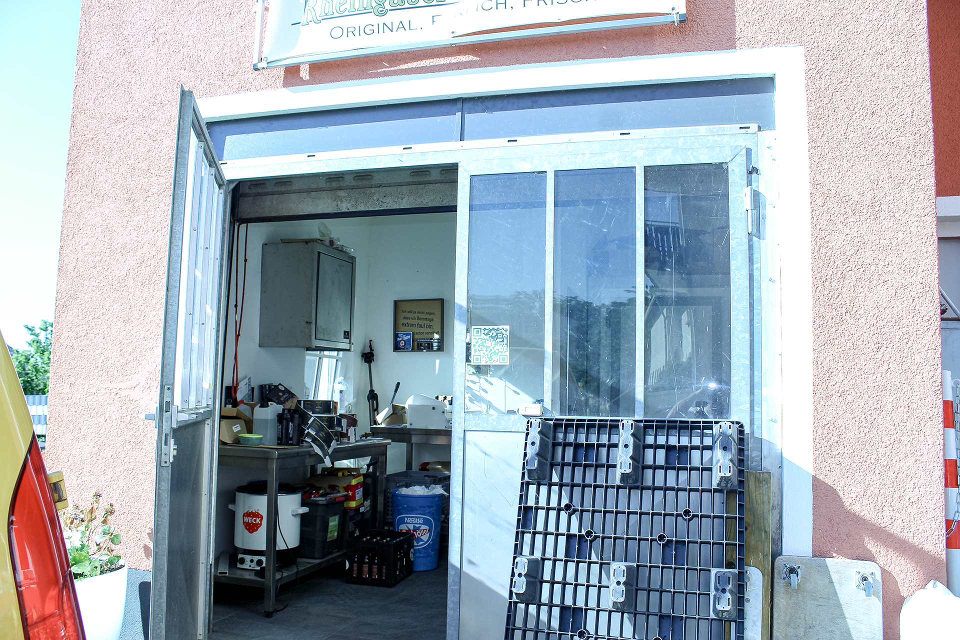 Innenansicht eines kleinen unfertigen Verkaufs- oder Arbeitsstandes mit Metallrahmen und gläserner Tür, im Hintergrund Regale mit Gegenständen, auf der Wand ein QR-Code und ein Zitat.
