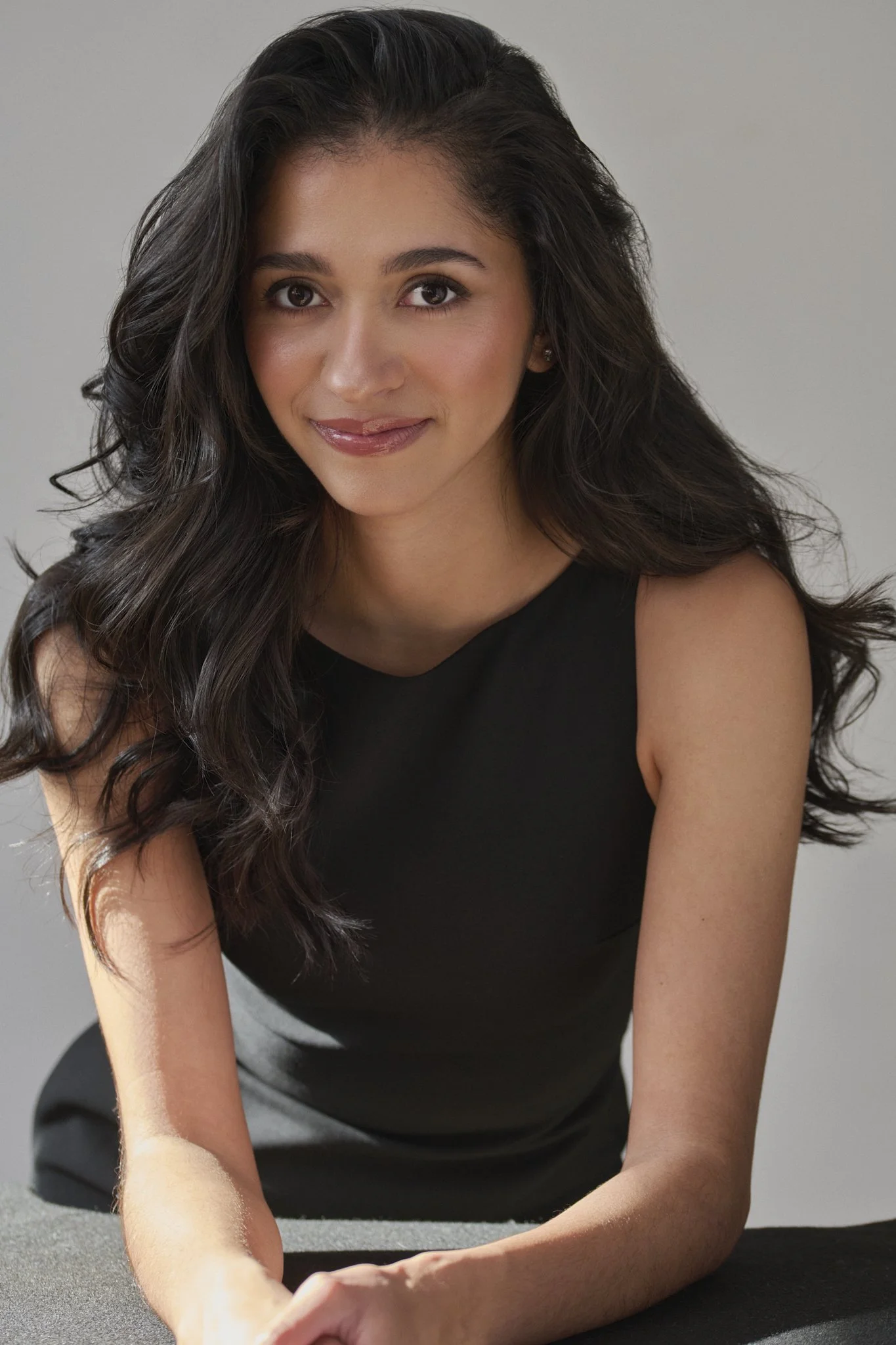 A woman with long, wavy dark hair, wearing a black sleeveless top, sitting at a table with her arms resting on it, smiling softly with a neutral gray background.