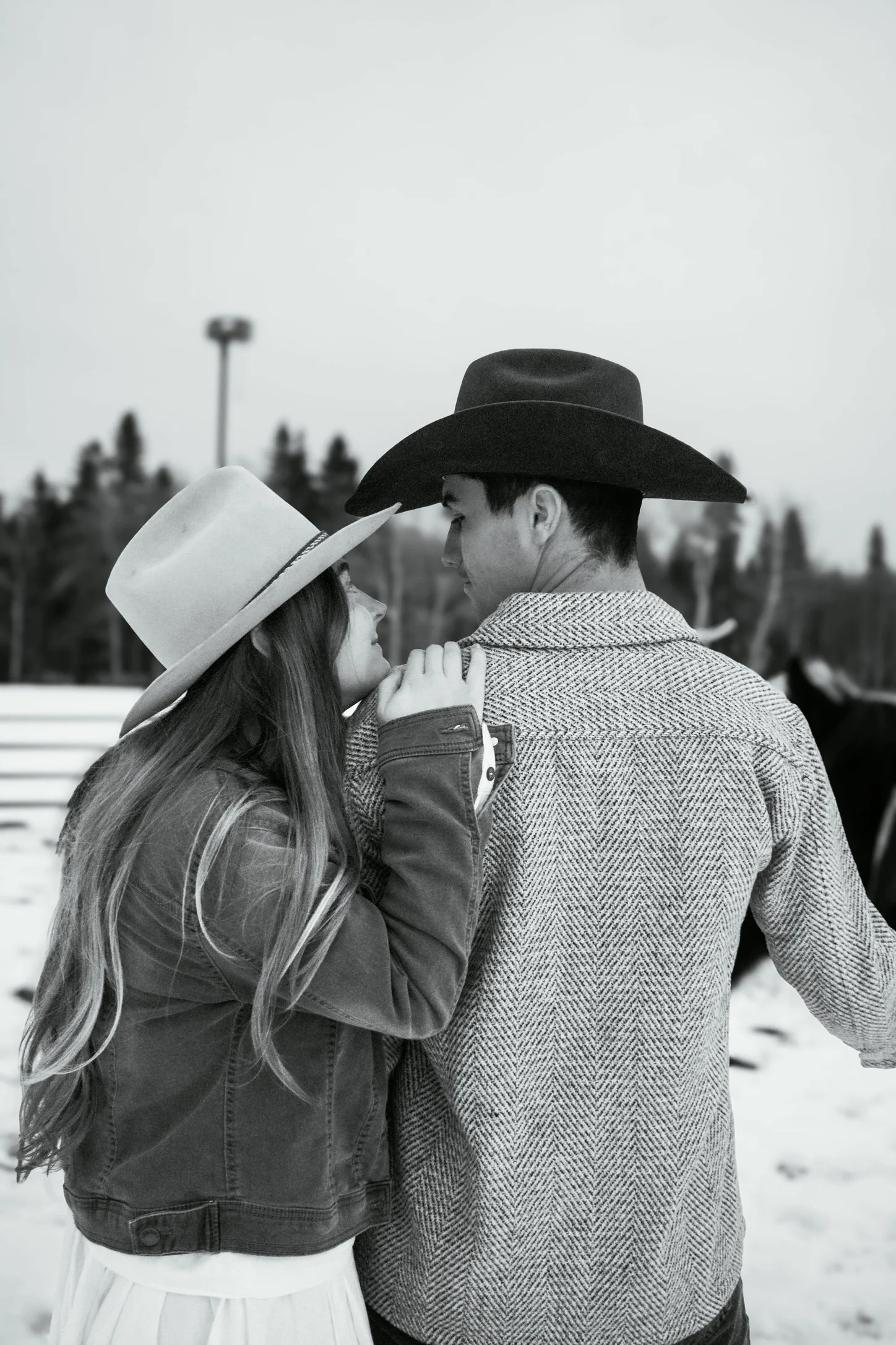 A black and white photo of a couple standing close and gazing into each other's eyes. The woman is wearing a wide-brimmed hat, and the man is wearing a large cowboy hat and a textured jacket. The background features blurred trees and a cloudy sky.