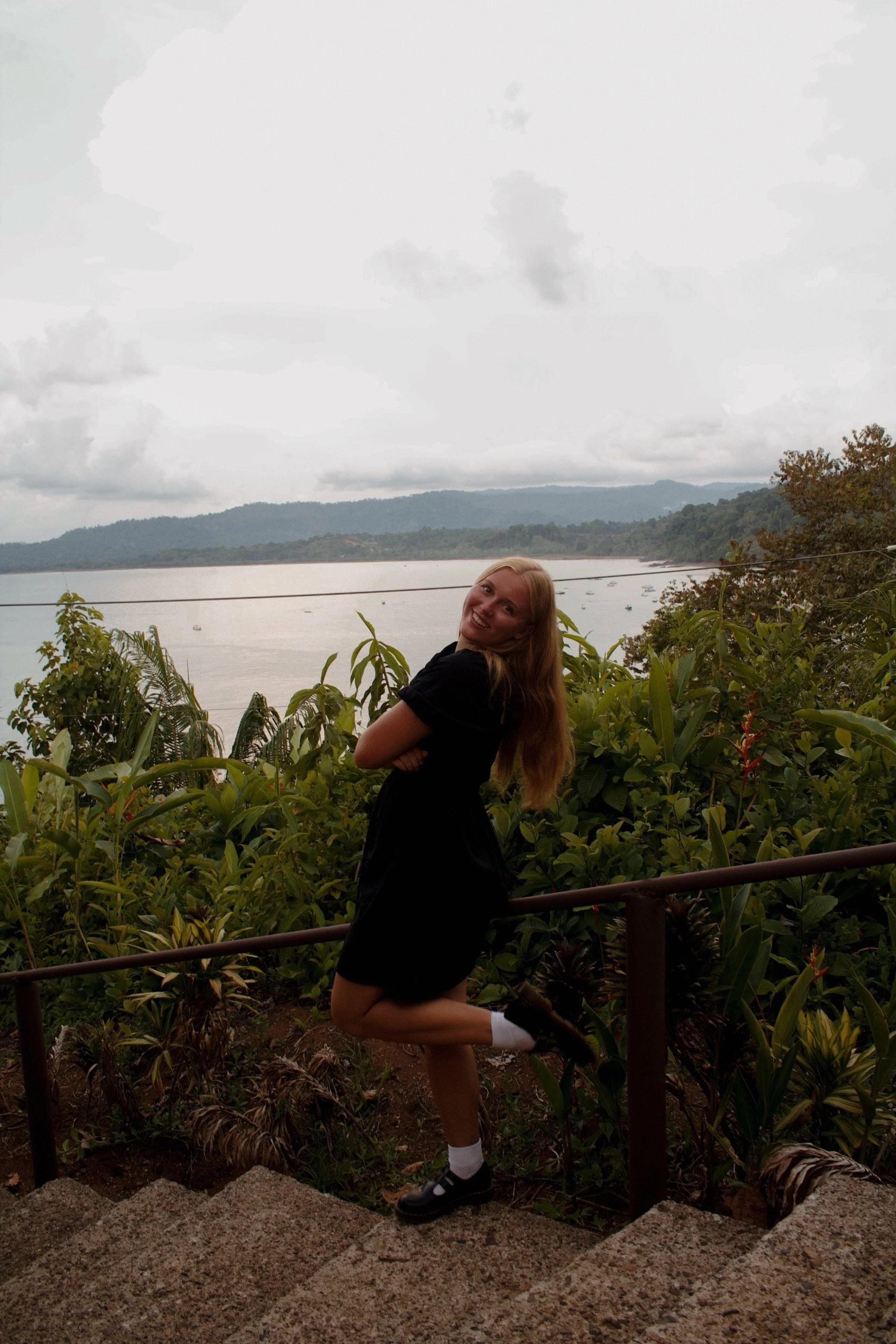 A young woman with long blonde hair stands on a pathway among lush green plants, smiling and posing with her arms crossed, in front of a body of water with boats and a mountainous landscape in the background on a cloudy day.