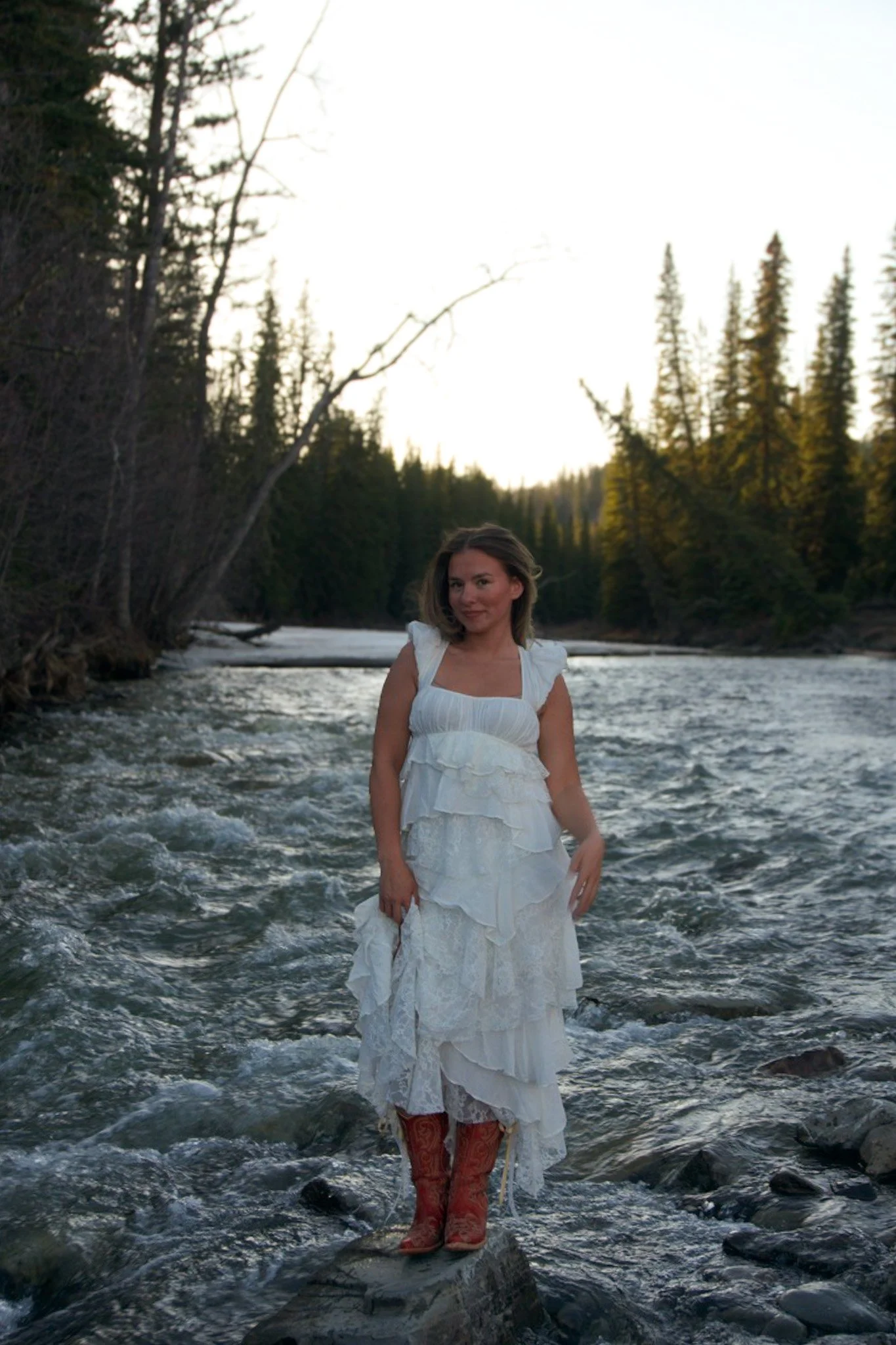 A woman in a white dress with ruffled layers stands knee-deep in a flowing river surrounded by trees, with a clear sky overhead.
