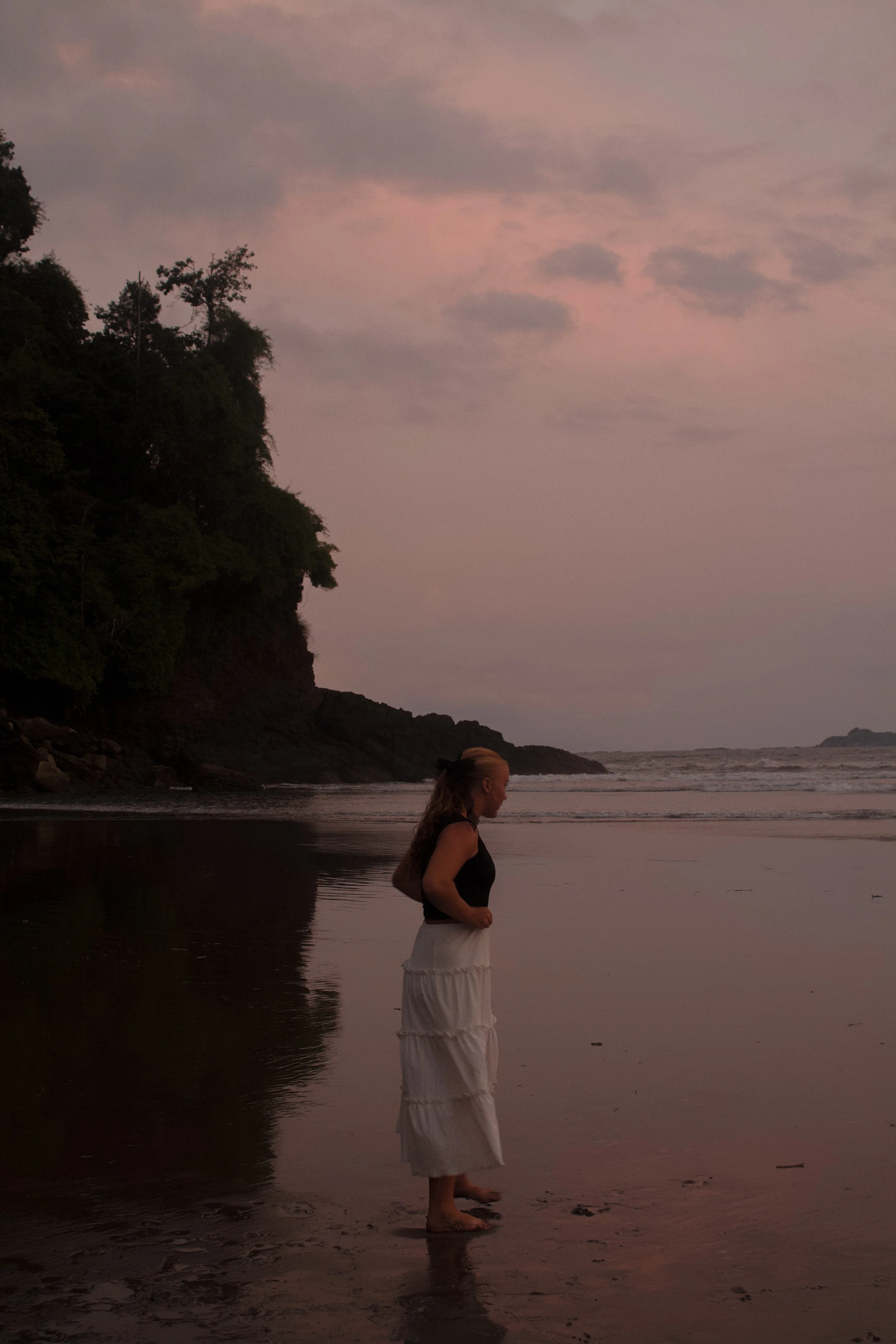 A woman standing barefoot on a sandy beach, facing the ocean with a rocky cliff covered in trees to her left, during dusk with pink and purple clouds in the sky.