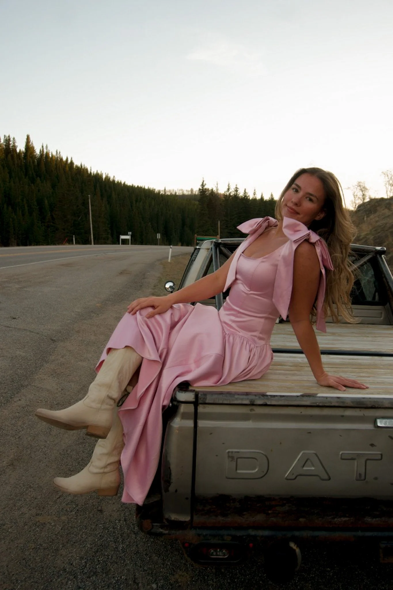 A woman in a pink dress and white cowboy boots sits on the tailgate of a pickup truck, smiling at the camera, with a forest and open road in the background during sunset.