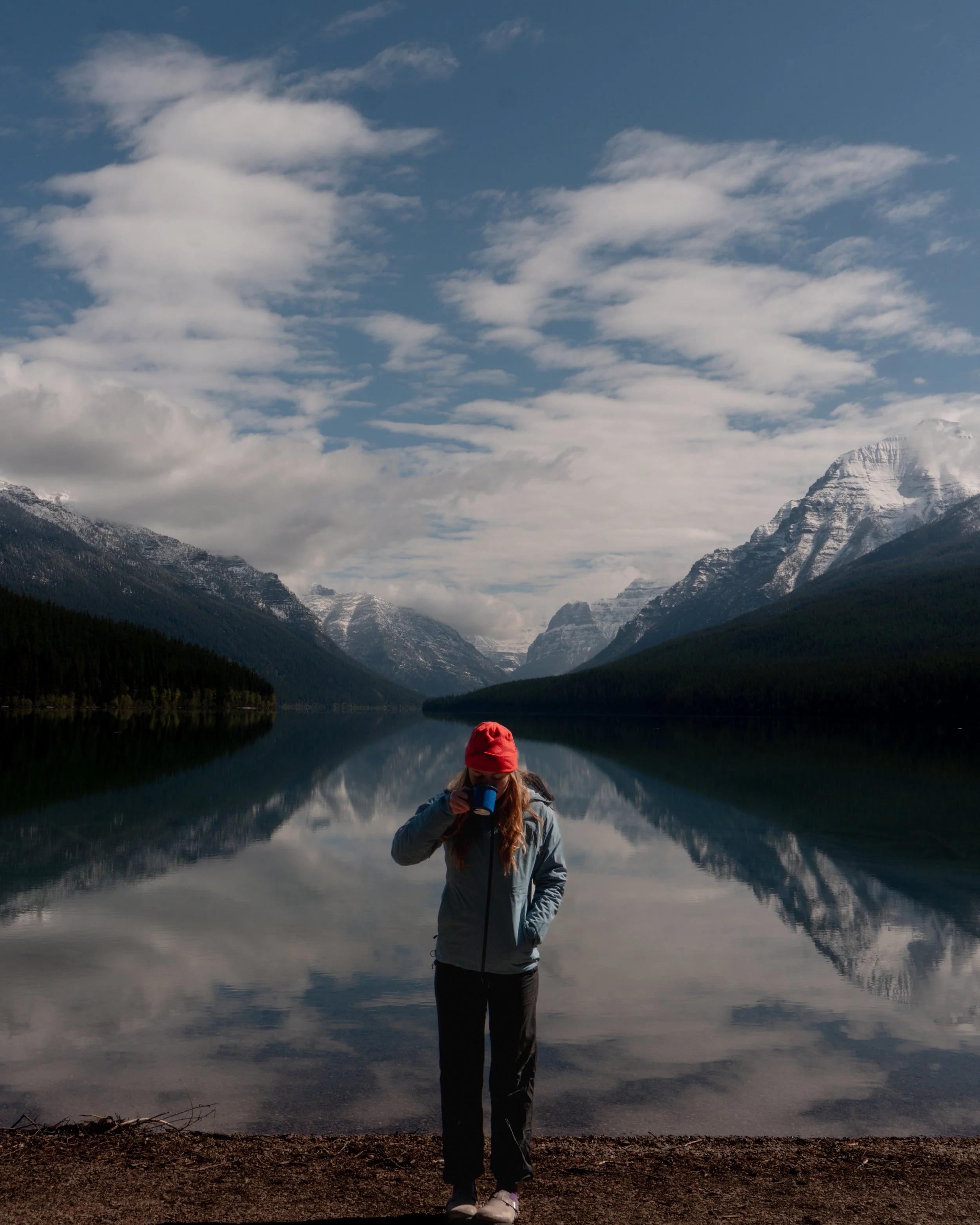 A person standing by a lake in a mountainous area, wearing outdoor gear and drinking from a mug, with snow-capped mountains and a partly cloudy sky reflected in the water.