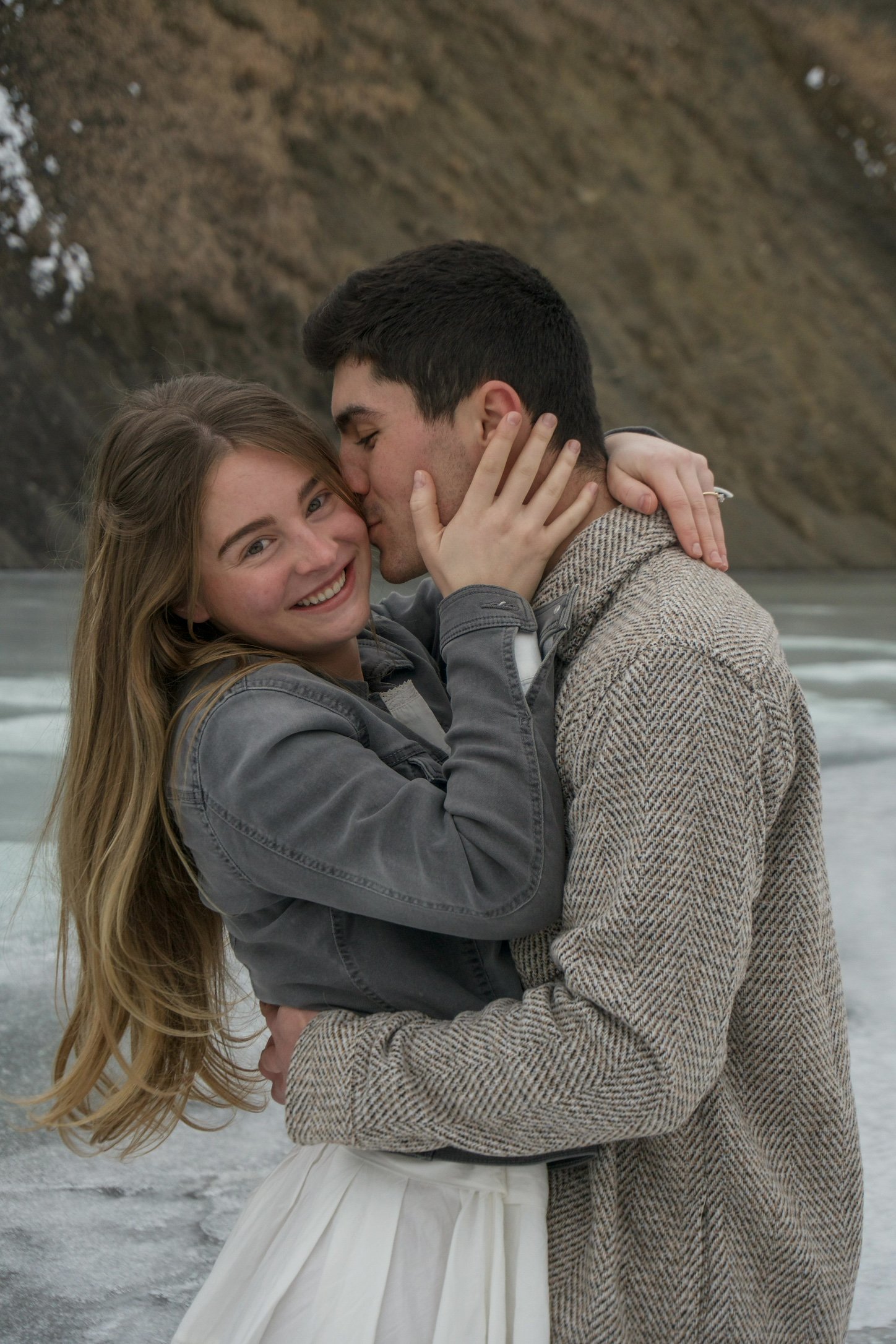 A smiling young woman with long, wavy hair is being embraced by a young man with short dark hair. The man is kissing her on the cheek while she is smiling and looking at the camera. They are outdoors near a body of water with a rocky cliff in the background.