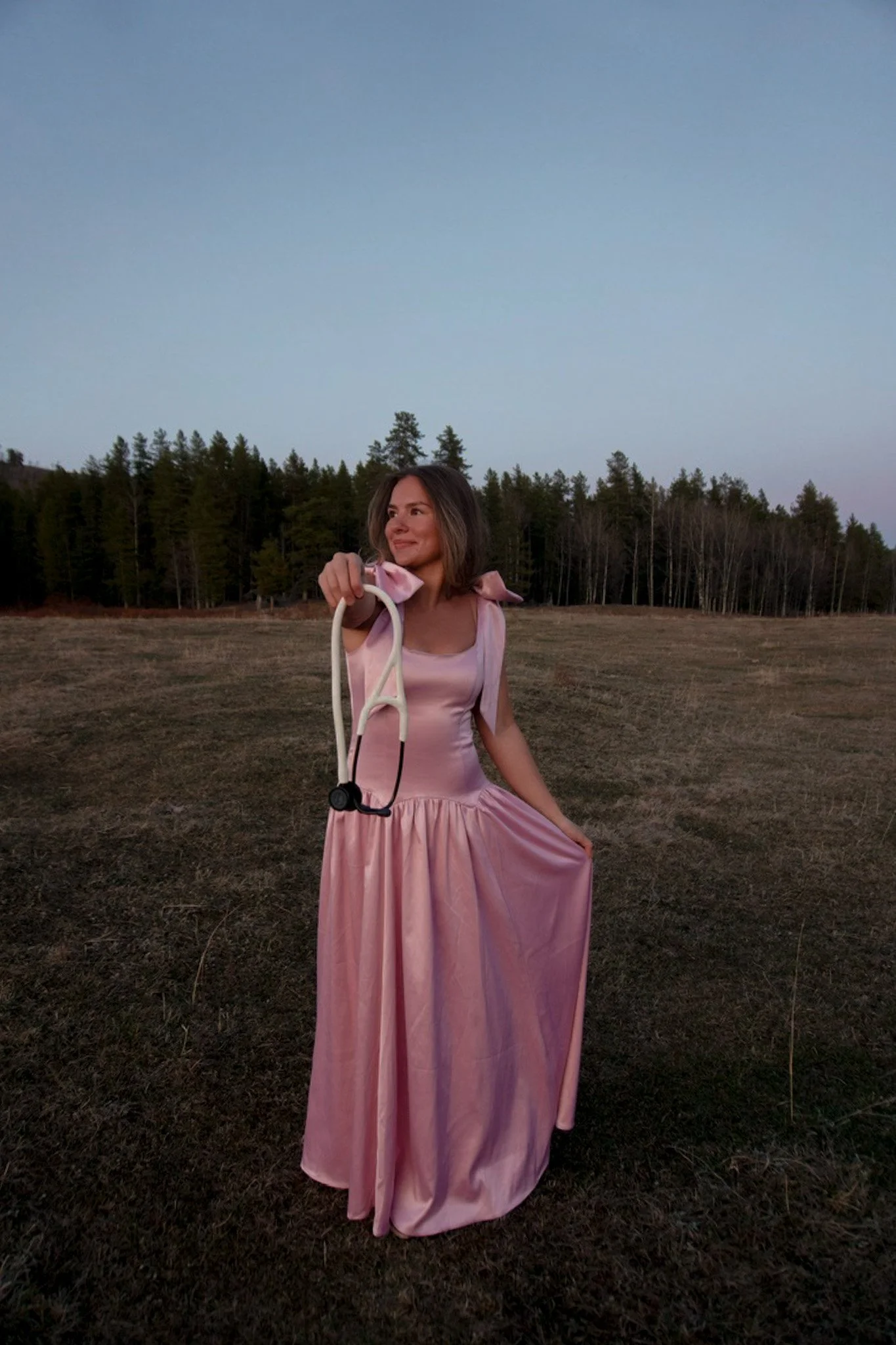 A woman in a pink dress holding a pair of white glasses with a black strap, standing outdoors in an open field with trees in the background during dusk or dawn.