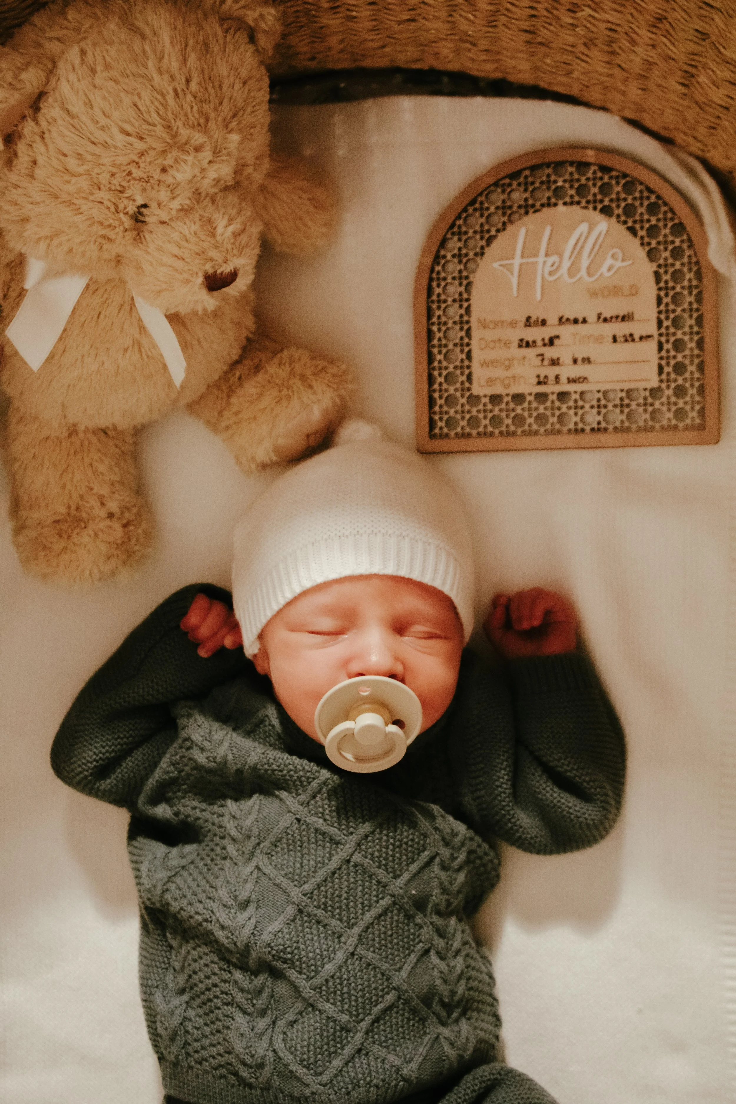 A baby lying on their back with a pacifier in their mouth, wearing a white knit hat and a dark gray sweater, next to a teddy bear and a wooden plaque with a birth announcement.