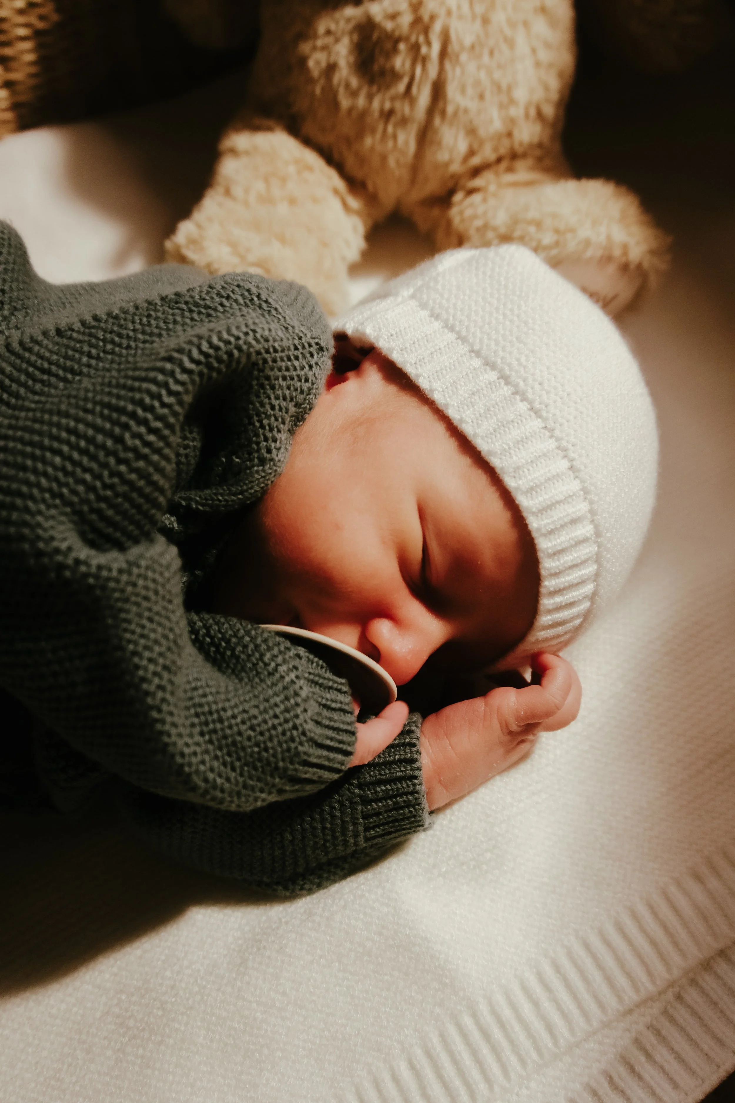 A sleeping newborn baby wearing a white knit hat and dark clothing, lying on a white textured blanket.