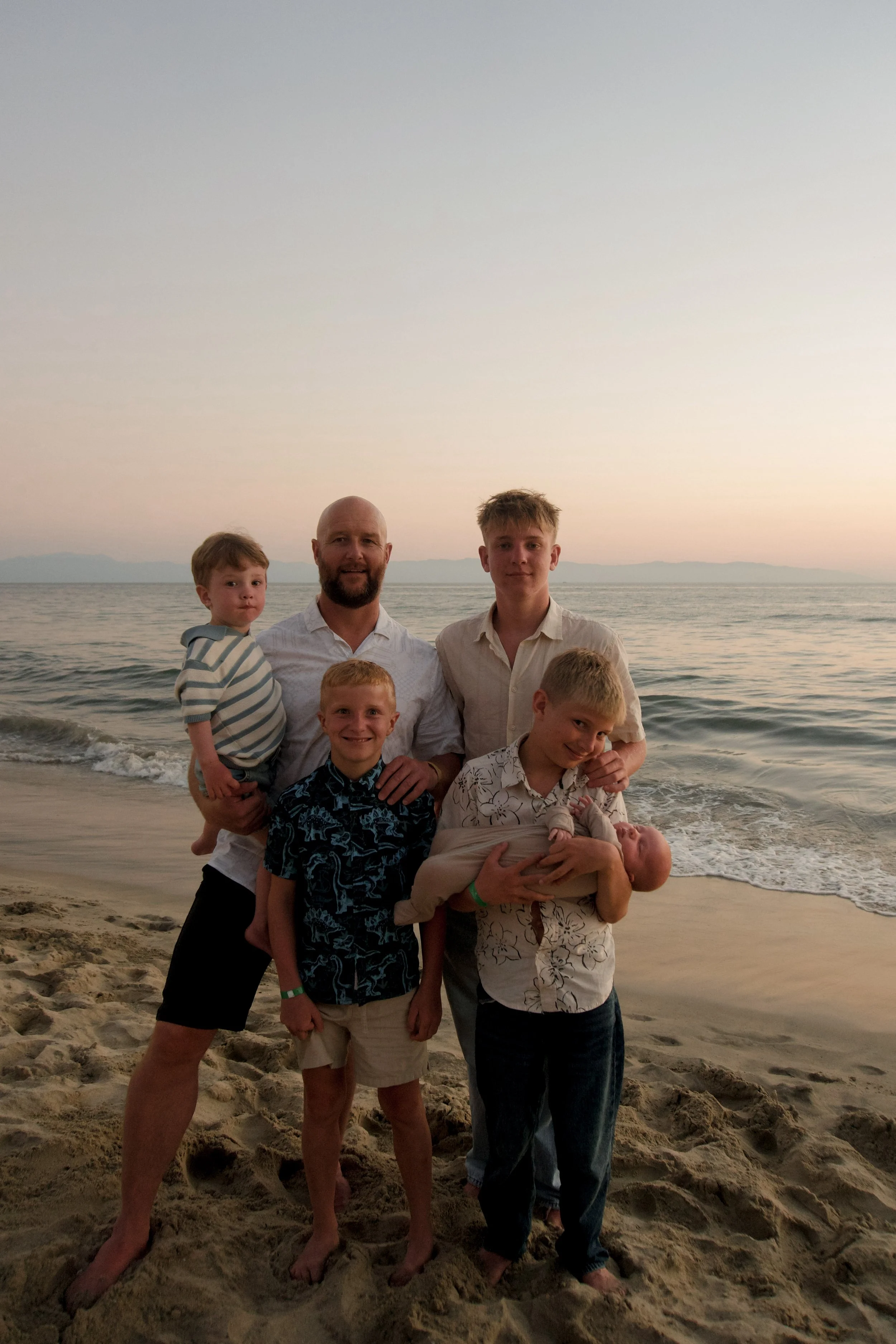 A group of seven people, including adults and children, stands on a sandy beach with the ocean and distant mountains in the background during sunset.