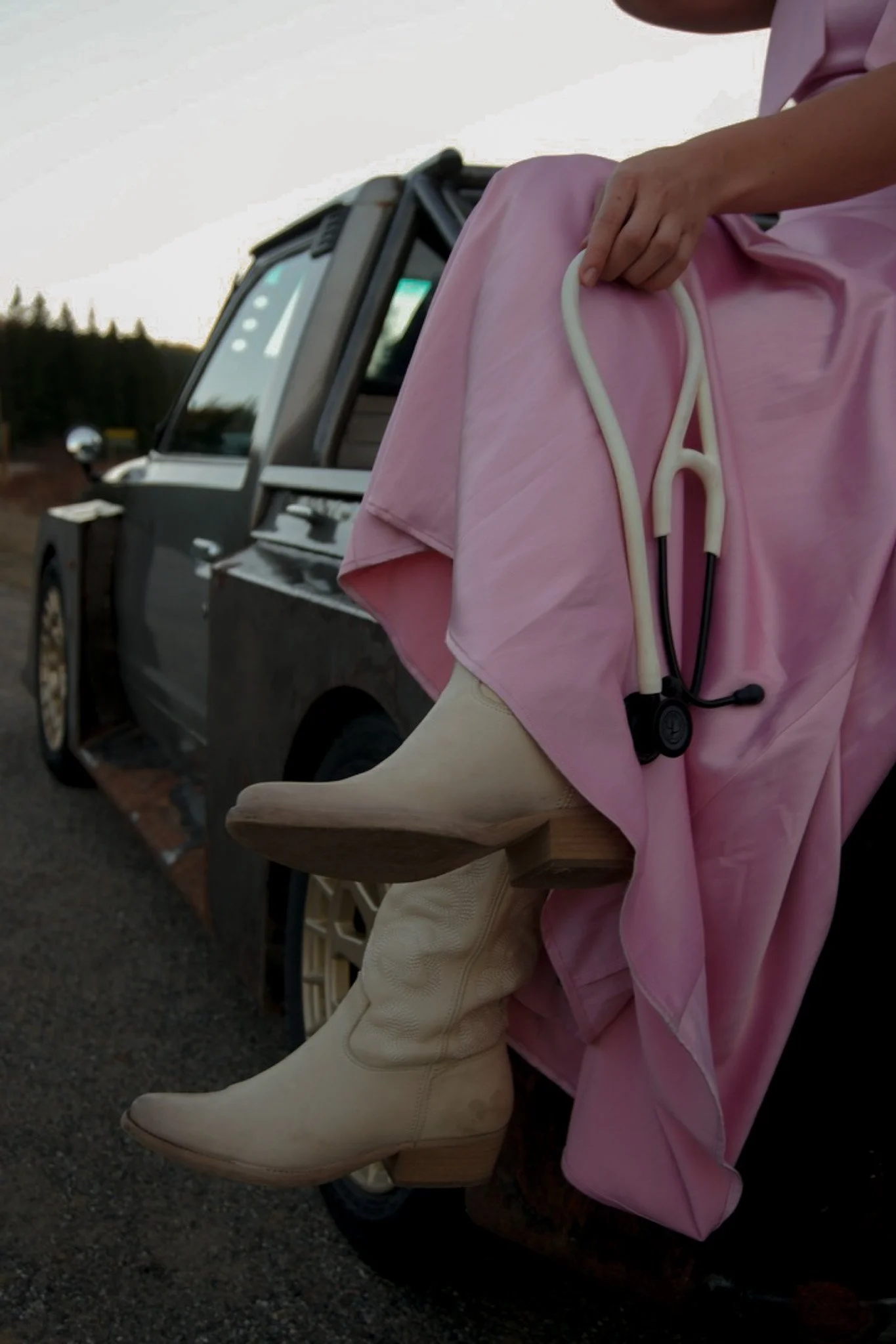 Close-up of a person's hand holding a white stethoscope on a pink fabric, with part of a beige boot visible and the door of a gray vehicle in the background.