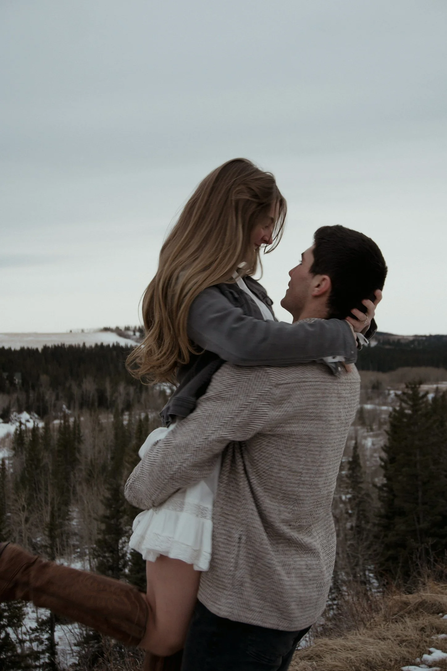 A man carrying a woman in his arms outdoors with a forested landscape and cloudy sky in the background.