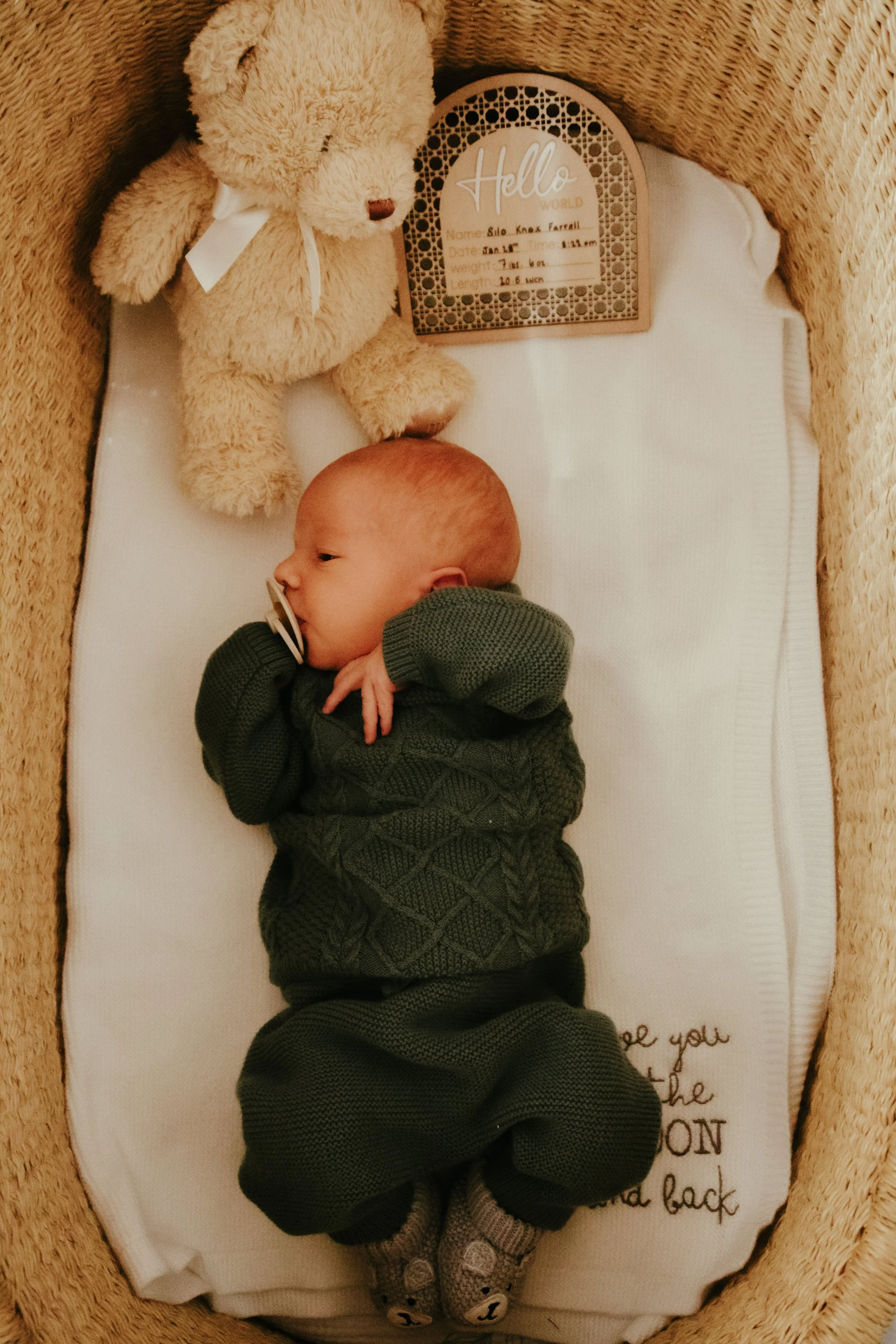 A newborn baby lying in a bassinet, wearing a dark green sweater and animal-themed socks, with a teddy bear and a decorative sign nearby.