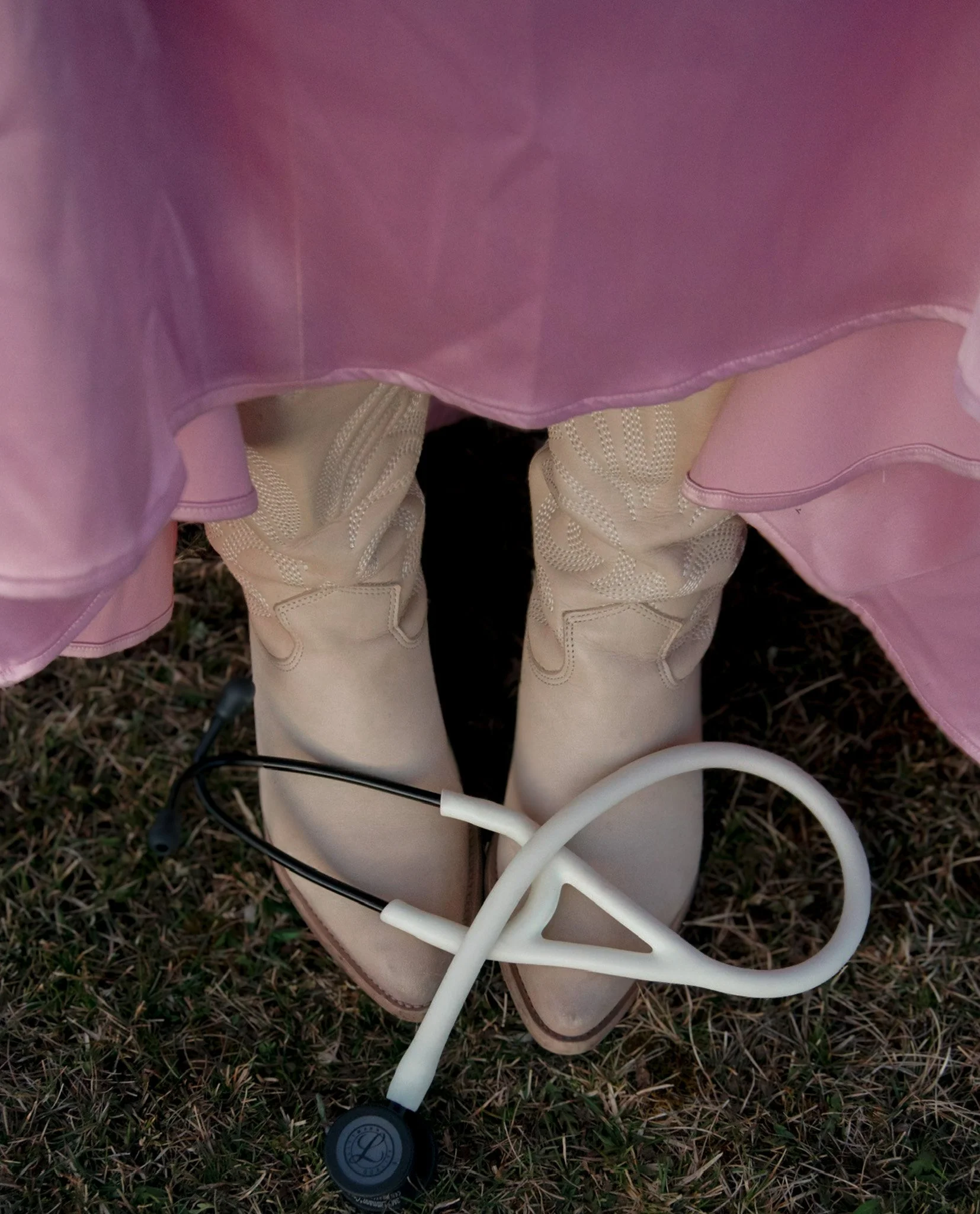 Close-up of a stethoscope placed on the ground with grass, a person wearing cream-colored pants, and a pink garment visible in the background.