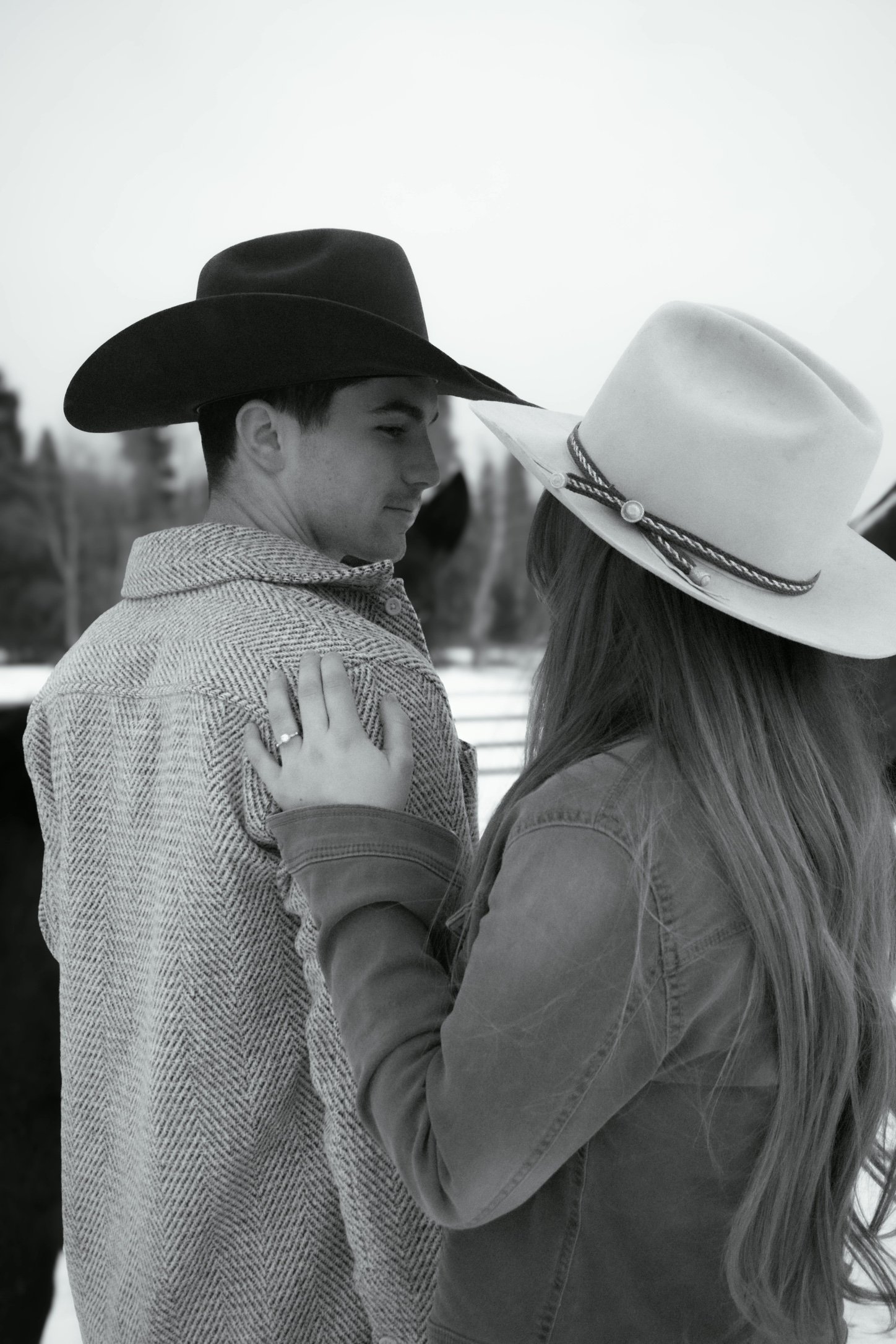 A black and white photograph of a young man and woman dancing closely outdoors in winter, both wearing cowboy hats. The man is in profile with a serious expression, wearing a patterned coat; the woman faces him with her hand on his shoulder, wearing a denim jacket and a wide-brimmed hat decorated with a band.