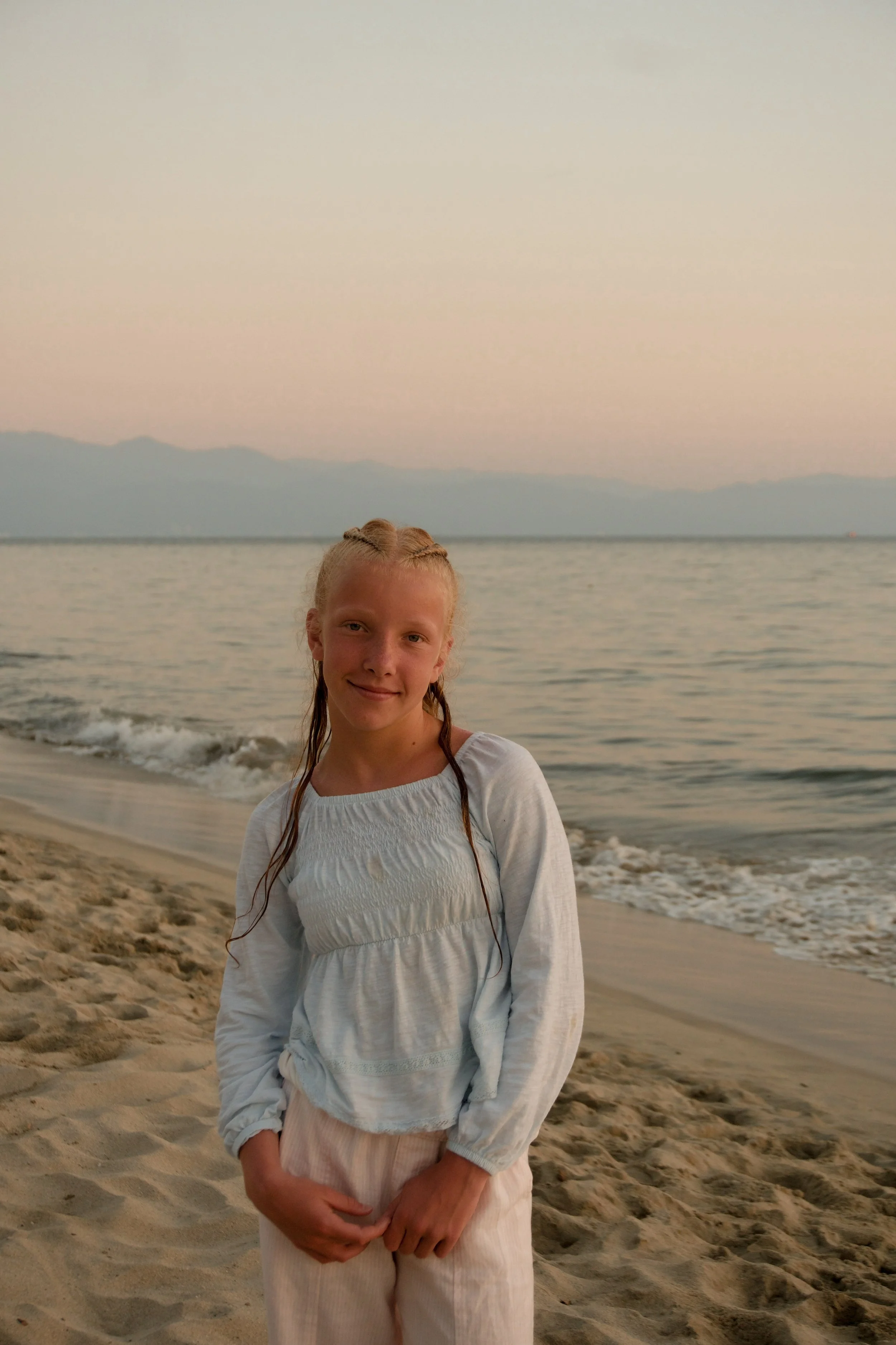 A young girl with braided hair, wearing a white long-sleeve top, standing on a sandy beach near the ocean at sunset, with mountains visible in the distance.
