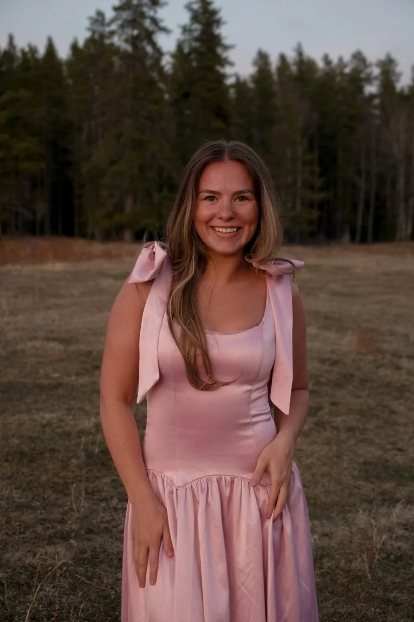 A young woman with long, wavy light brown hair smiling outdoors during dusk or early evening, wearing a pink sleeveless dress with ruffled shoulders, standing in a grassy field with tall trees in the background.