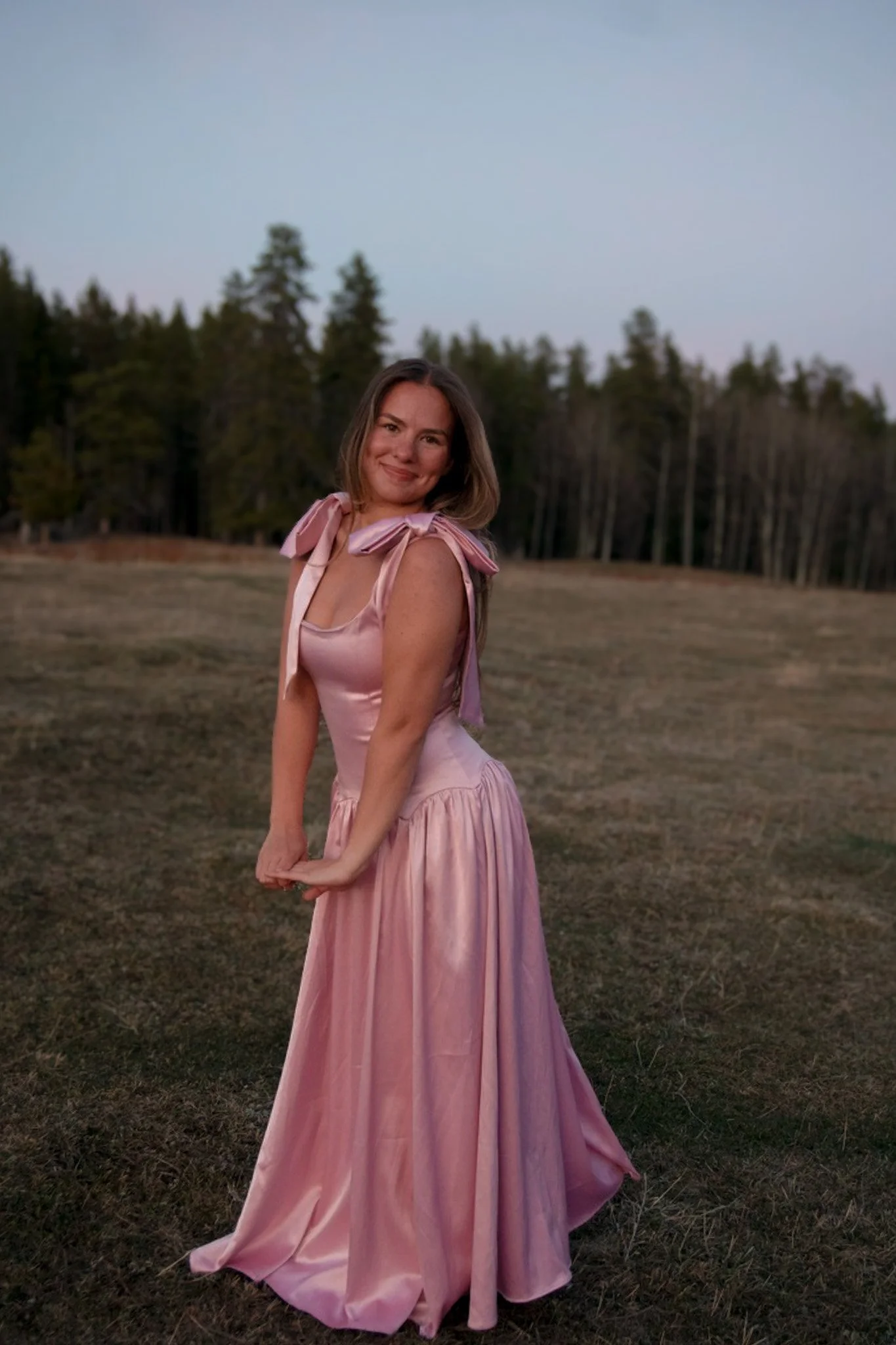 A woman in a flowing pink dress with bow details on the shoulders standing on a grassy field with a forest in the background during dusk, smiling at the camera.