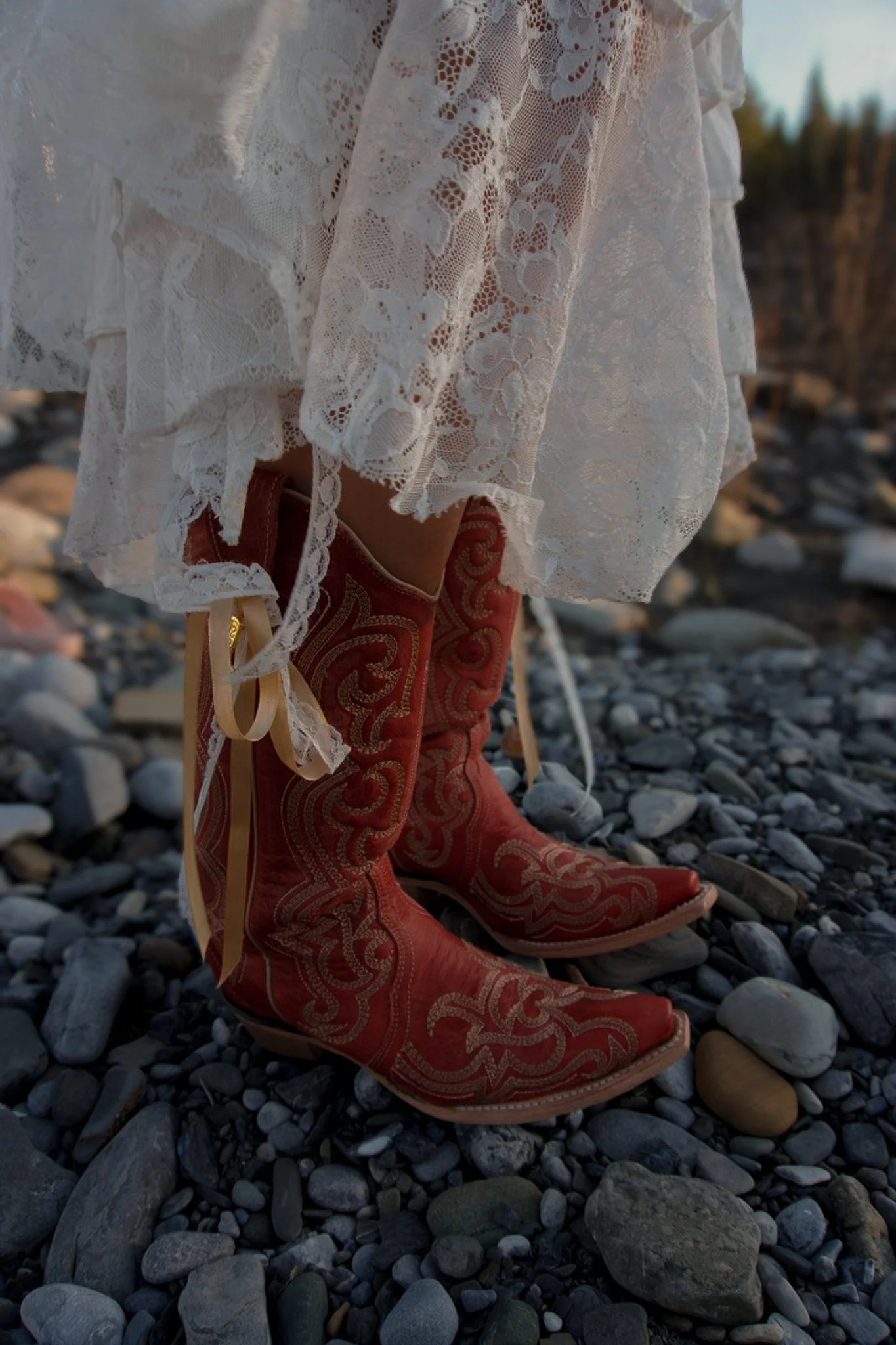 Close-up of a person's lower legs wearing red cowboy boots with gold embroidery, positioned on a rocky surface, with a white lace dress flowing around the boots.