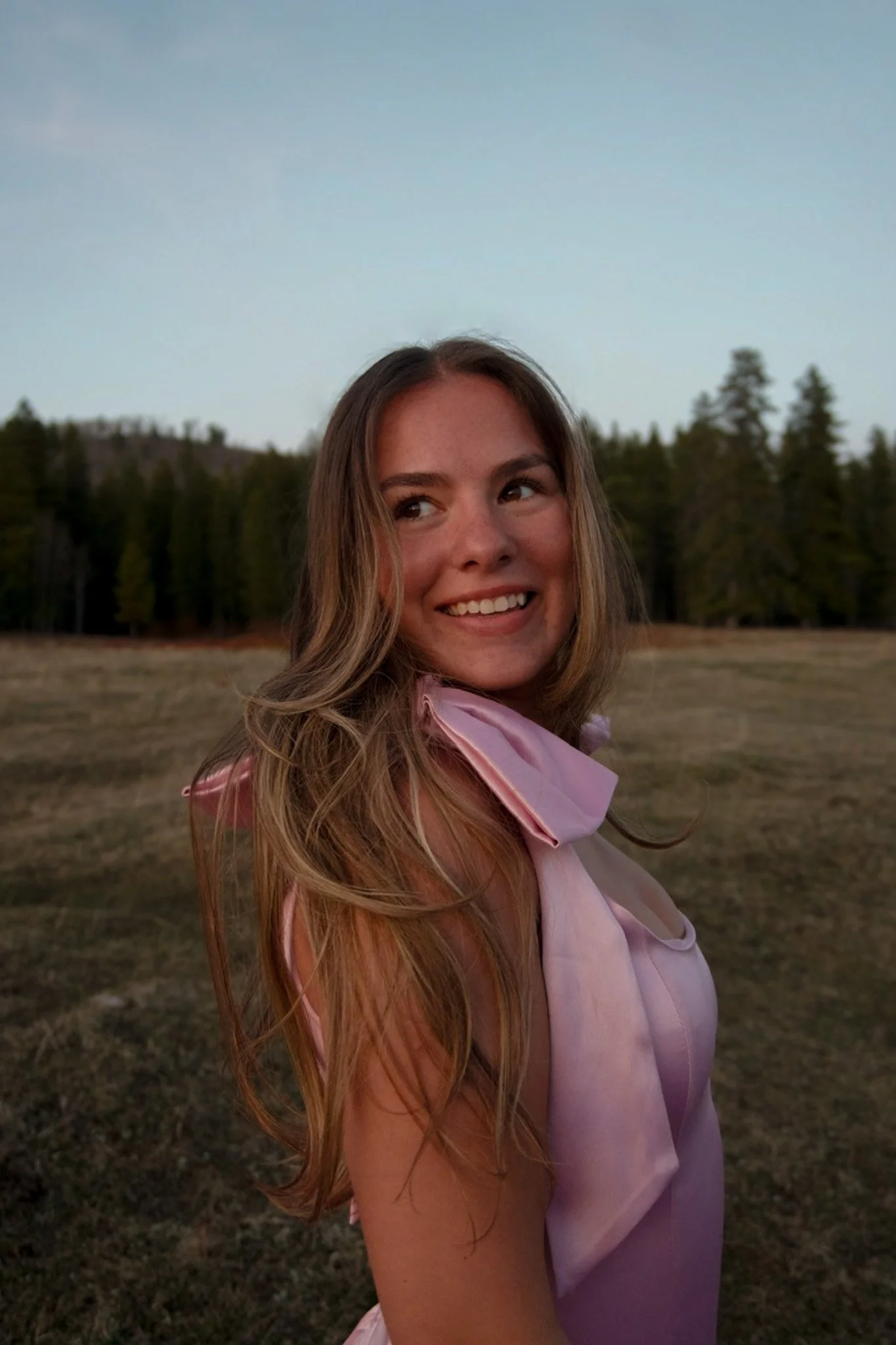 A young woman with long wavy hair smiling outdoors at sunset, with a background of trees and an open field.