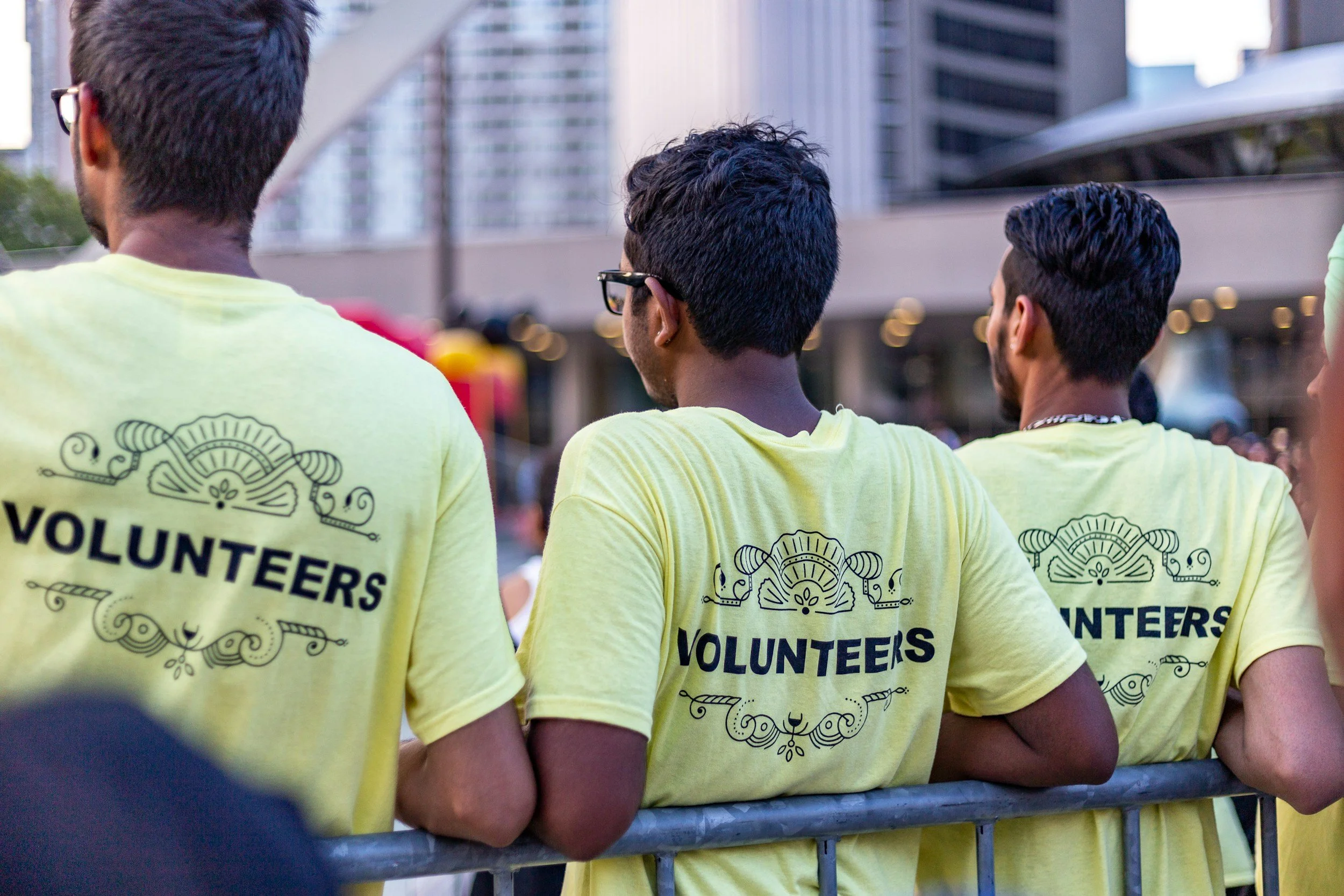 Three volunteers wearing yellow T-shirts standing behind a metal barrier in an urban environment.