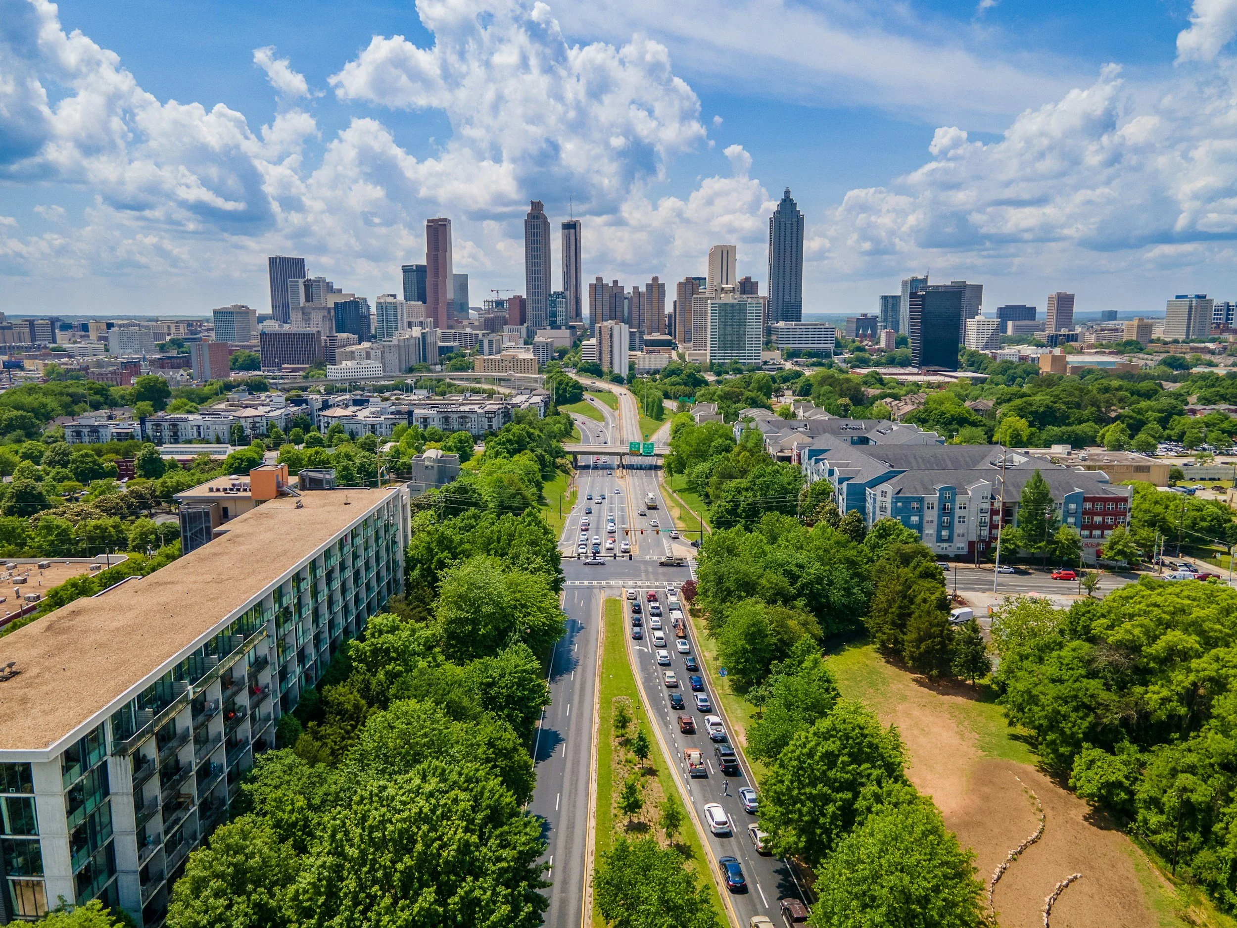 A city skyline with modern skyscrapers and buildings, a busy multi-lane road with cars, and lush green trees, under a partly cloudy blue sky.