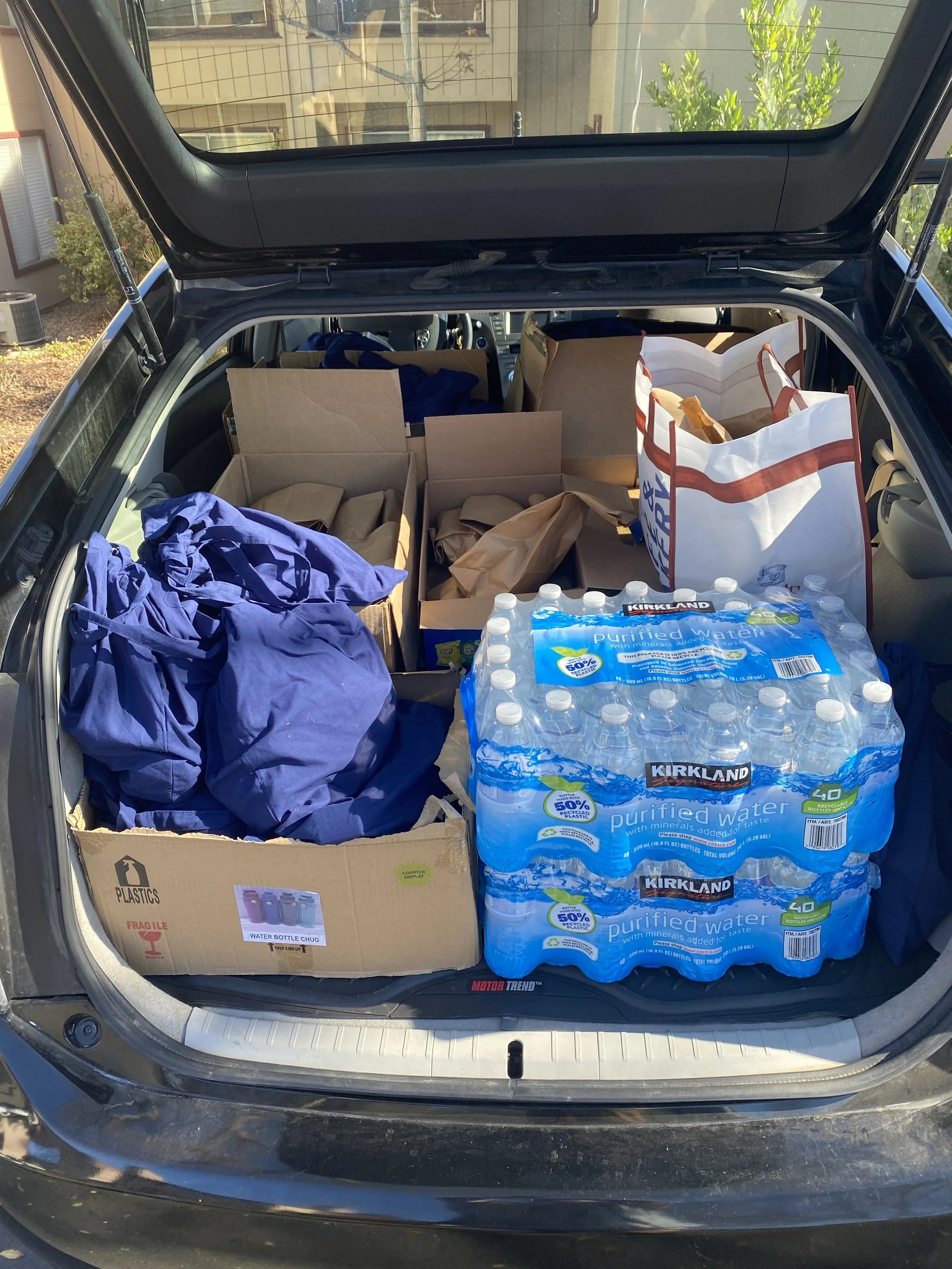 Open car trunk filled with cardboard boxes, paper bags, a pack of bottled water, and various items, with a house and trees visible in the background.