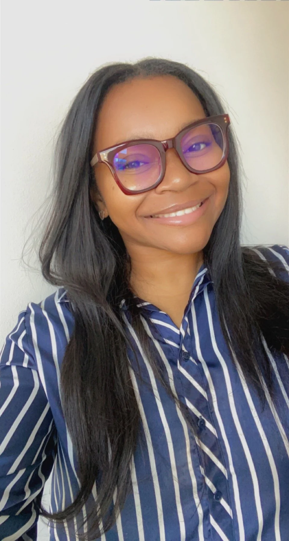 A smiling woman with long black hair, glasses, and a striped blue and white shirt, taking a selfie against a plain white wall.