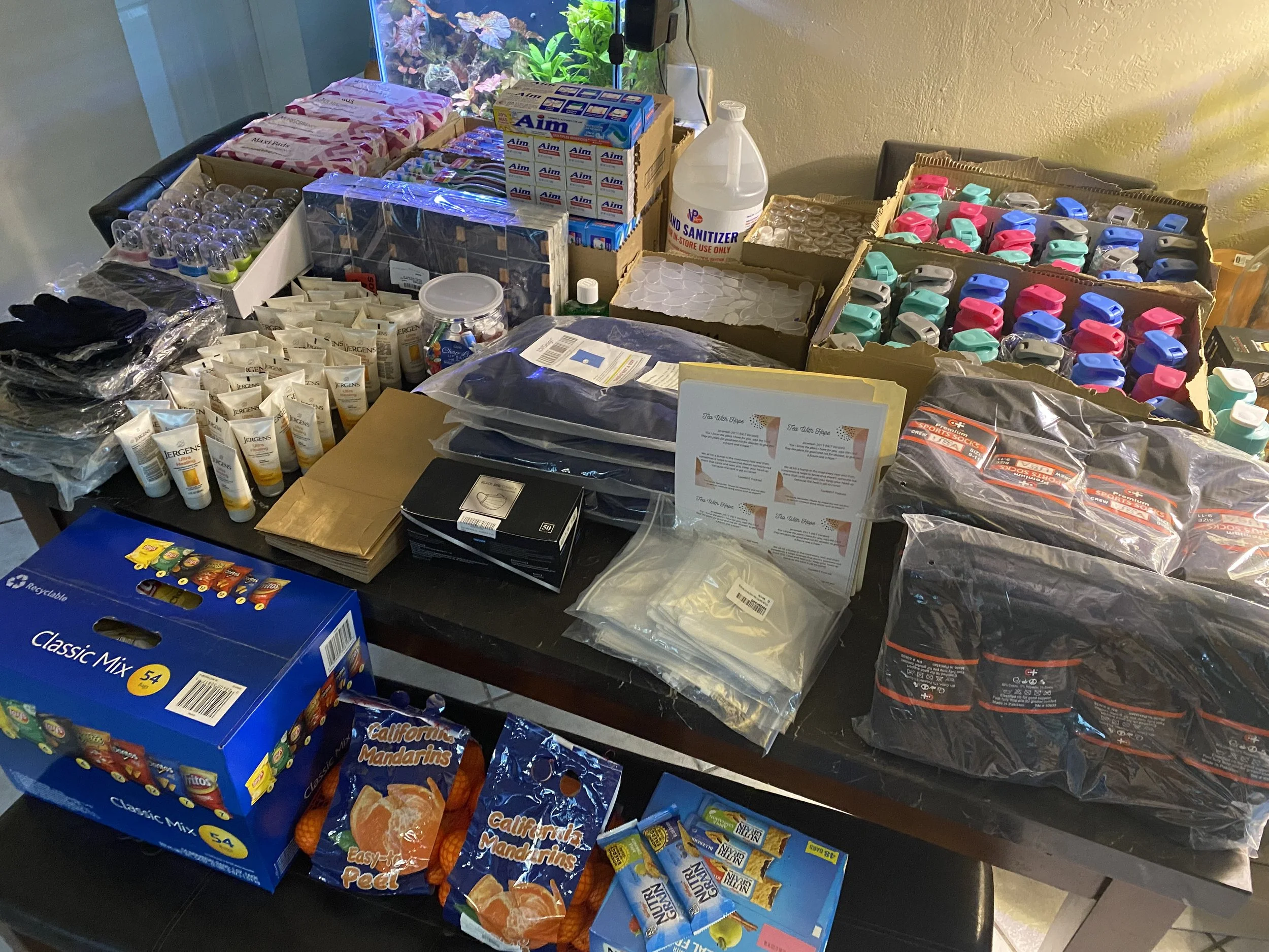 Table filled with various supplies including snack packs, boxes of tissues, bottles of sanitizer, colorful clips, small plastic containers, and other miscellaneous items.