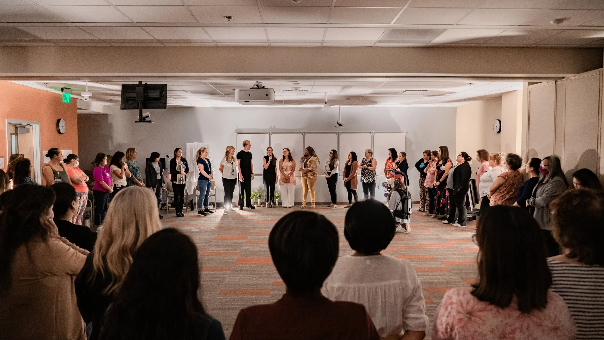 People gathered in a meeting room, standing in a semi-circle, with one person speaking into a microphone, and others listening.