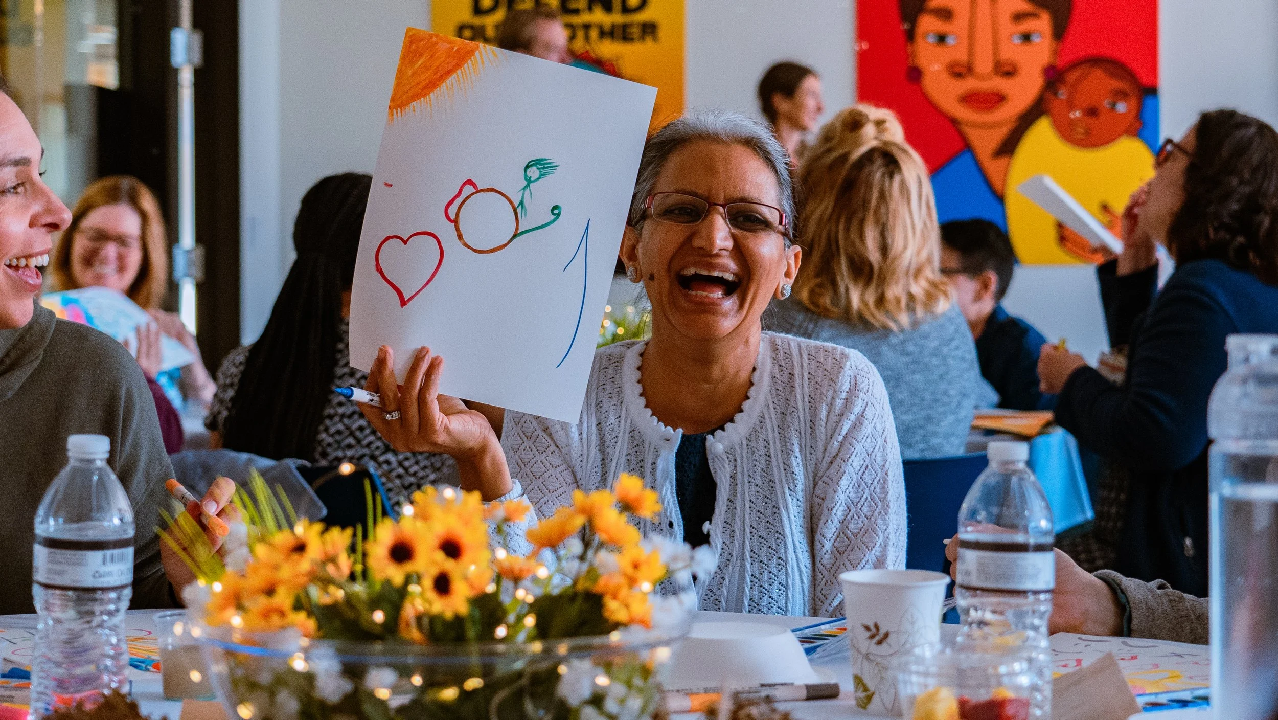 A woman smiling and holding up a drawing with a sun and hearts at a lively indoor gathering with people sitting at tables, decorated with flowers and bottled water.