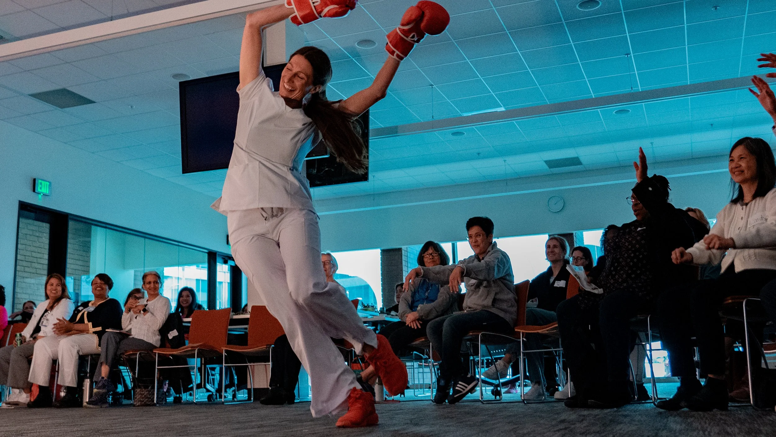 A woman in white pants and red boots dances enthusiastically in front of an audience seated in chairs indoors, with several people smiling and watching her.