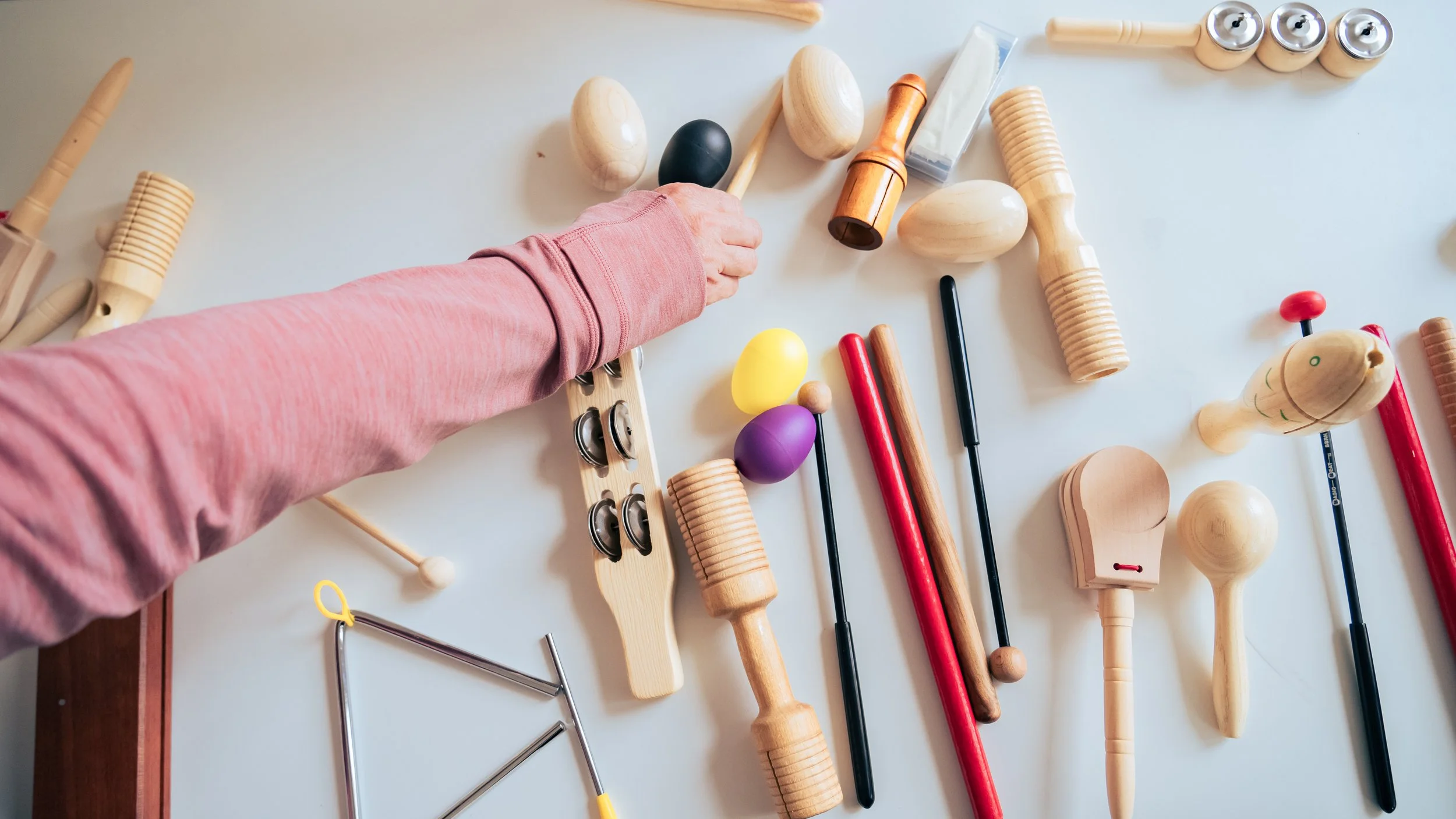 A person in a pink long sleeve shirt reaching for or placing a wooden musical instrument amidst various wooden and plastic musical toys and instruments on a light-colored surface.