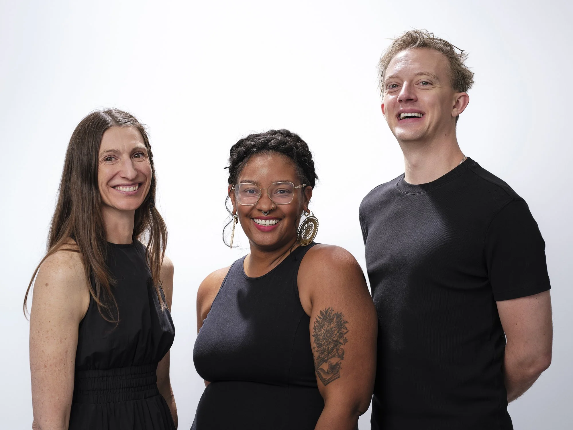 A diverse group of three women, standing close together, smiling at the camera against a plain white background.