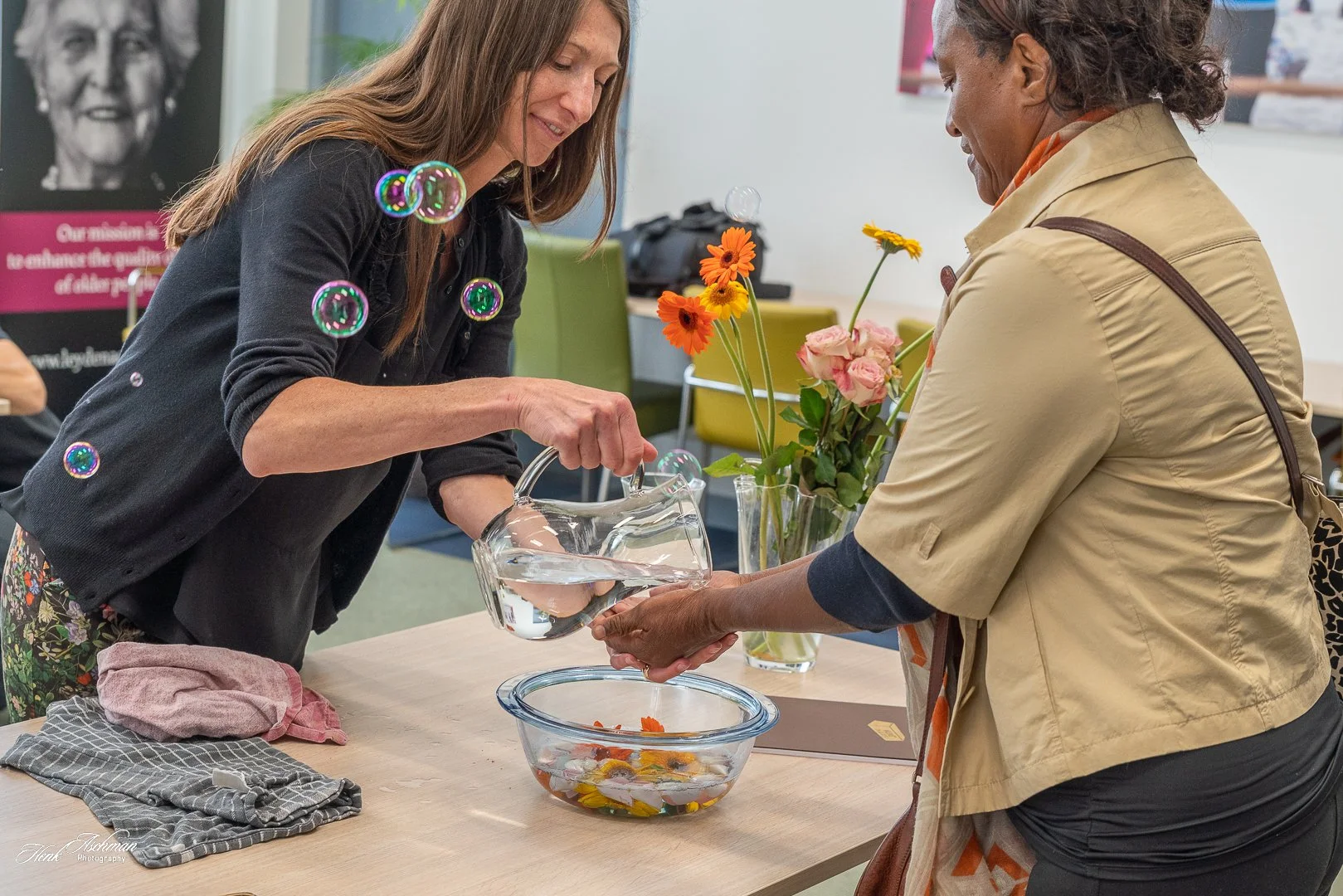 Two women exchanging flowers and water in a glass pitcher at a table with a vase of colorful flowers, in an indoor setting.