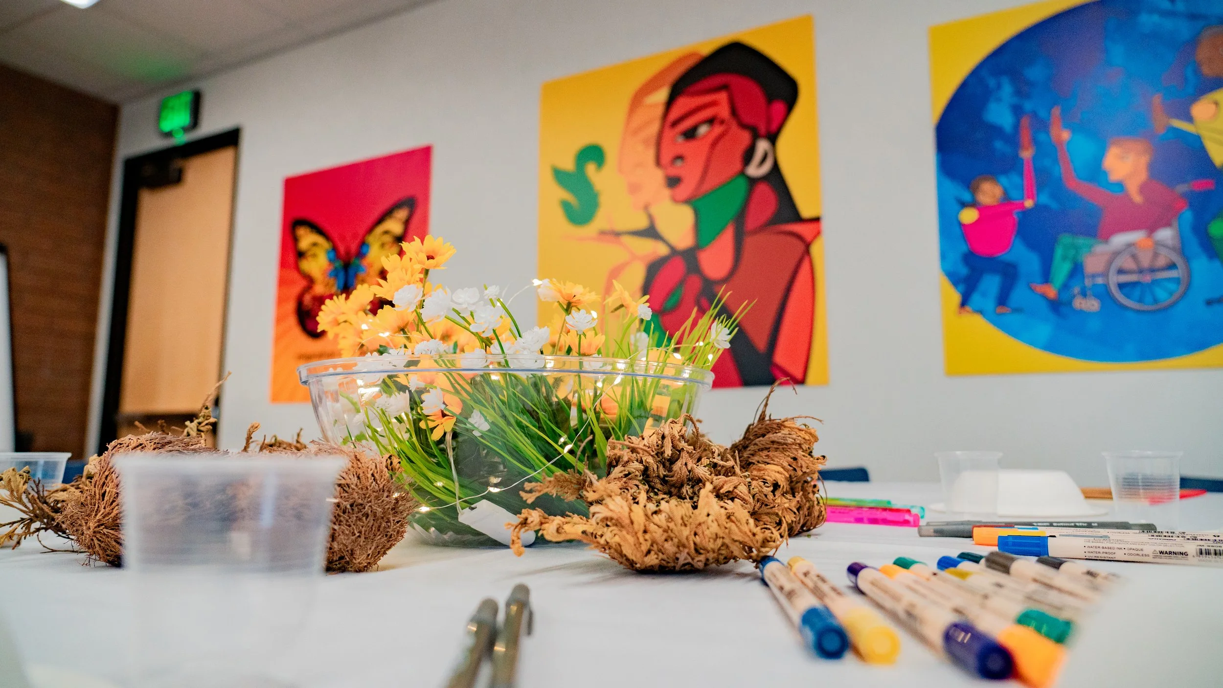 A table with art supplies, including markers and a centerpiece of yellow and white flowers in a clear bowl, with colorful paintings on the wall in the background.