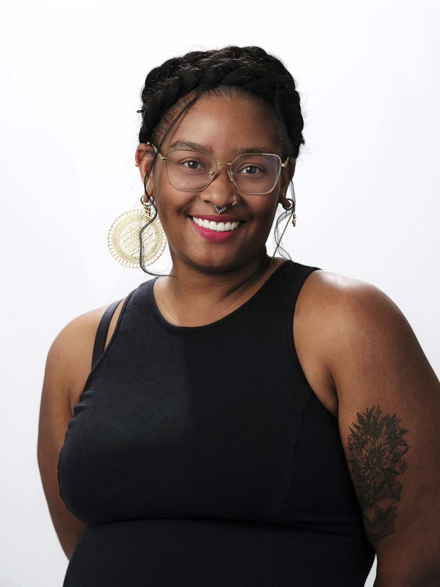 Portrait of a smiling woman with glasses, large gold earrings, and a tattoo on her upper arm, against a white background.