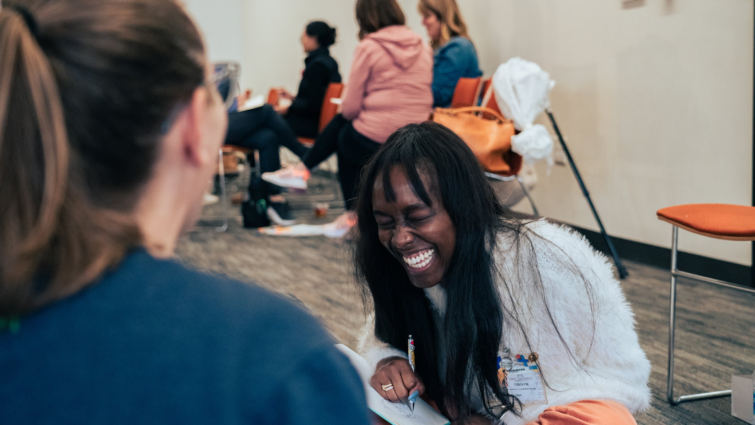 A woman with long dark hair laughing joyfully as she interacts with another person, with several people seated on chairs in the background, in an indoor setting.