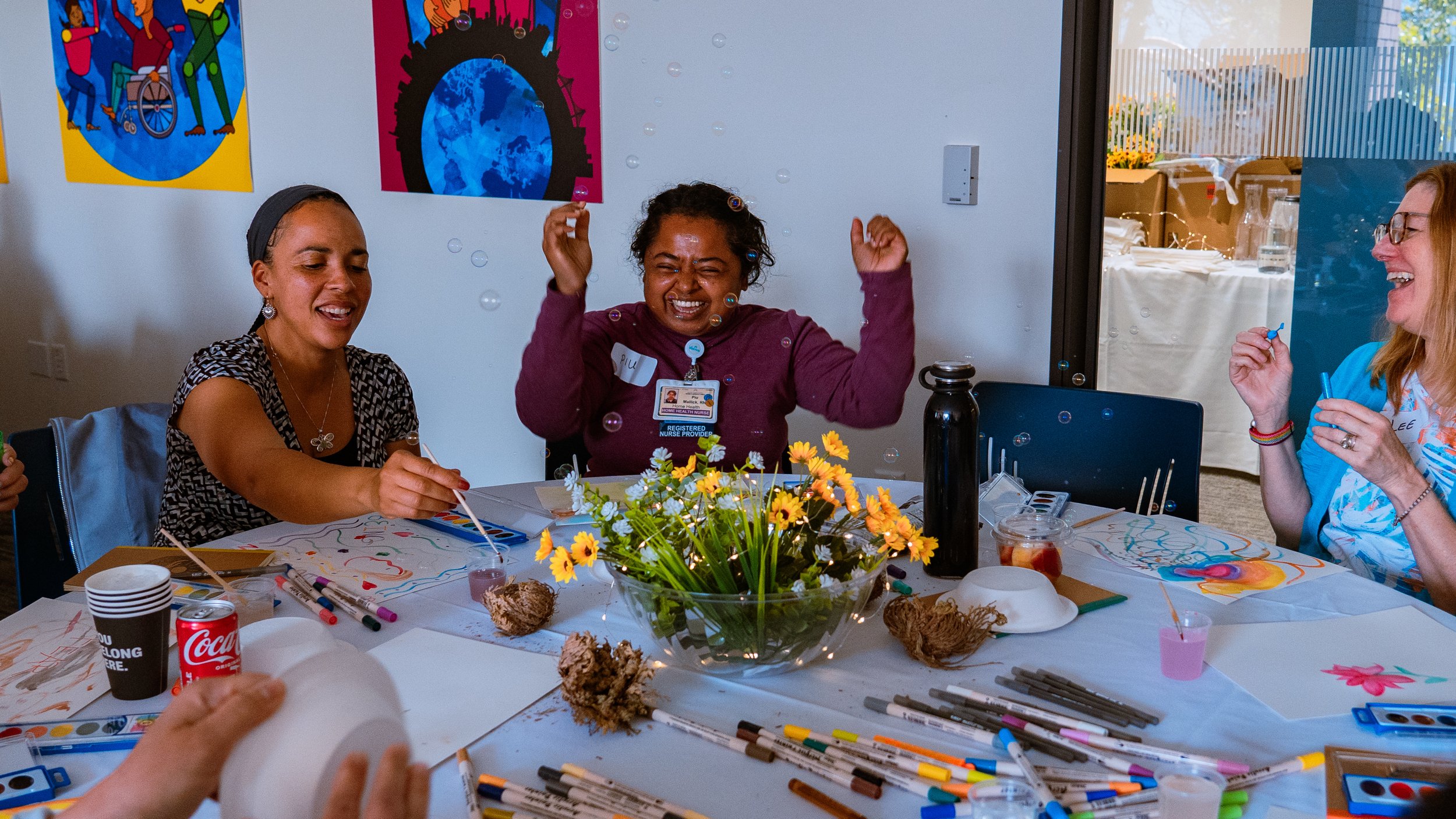 People sitting around a table with art supplies, drawing and painting, young woman in the middle with curly hair and a name tag, laughing and raising her hands, surrounded by colorful artwork and flowers on the table.