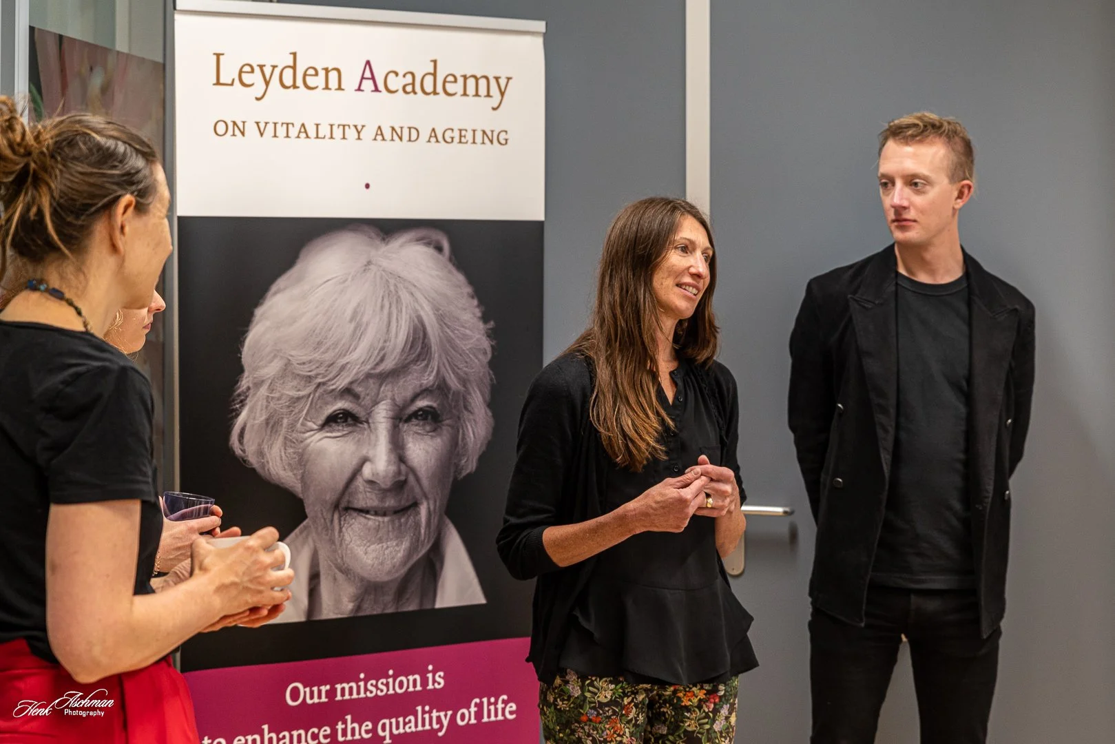 Group of people standing and talking at an event with a banner behind them that reads 'Leyden Academy on Vitality and Ageing' and features a black and white photo of an elderly woman. The women are holding drinks and engaging in conversation.