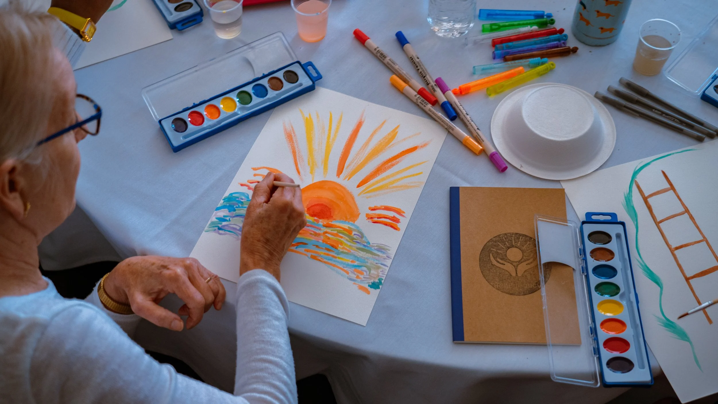 A person painting a colorful sunrise or sunset landscape on paper using various watercolor paints and brushes, surrounded by art supplies on a table.