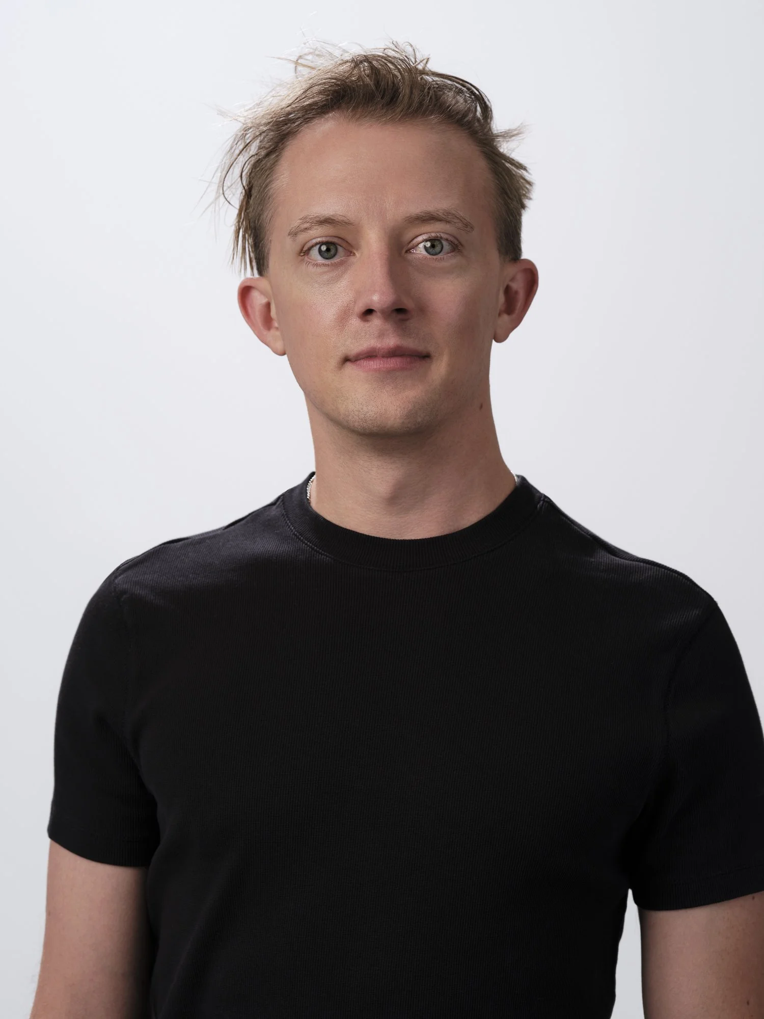 A young man with light skin, blue eyes, and short, messy, light brown hair, wearing a black t-shirt, standing against a plain light gray background.